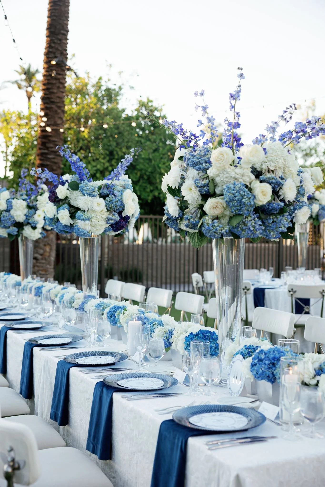 Outdoor event setup with long dinner tables decorated with blue, white, and purple flowers. Elegant table settings include navy blue napkins, clear glassware, and plates. Tall floral centerpieces with blue and white flowers are visible, with a palm tree and string lights in the background.