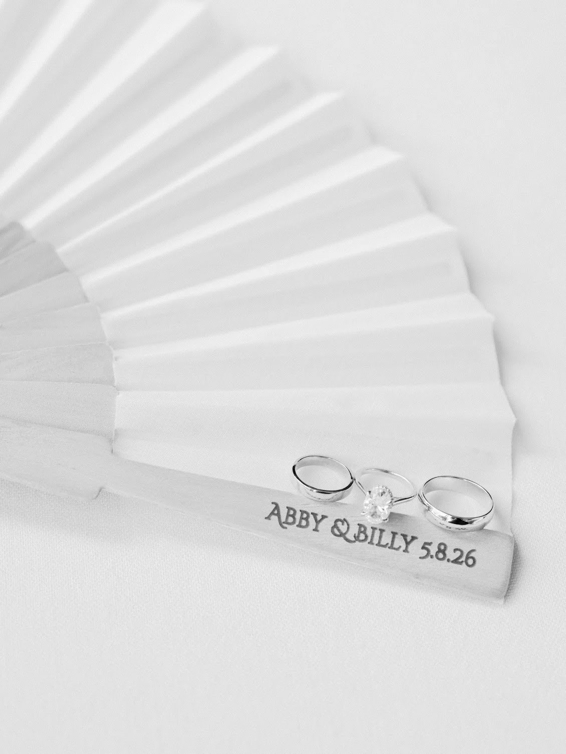 Wedding rings and a small plaque with the inscription "ABBY & BILLY 5.8.26" on a white fan