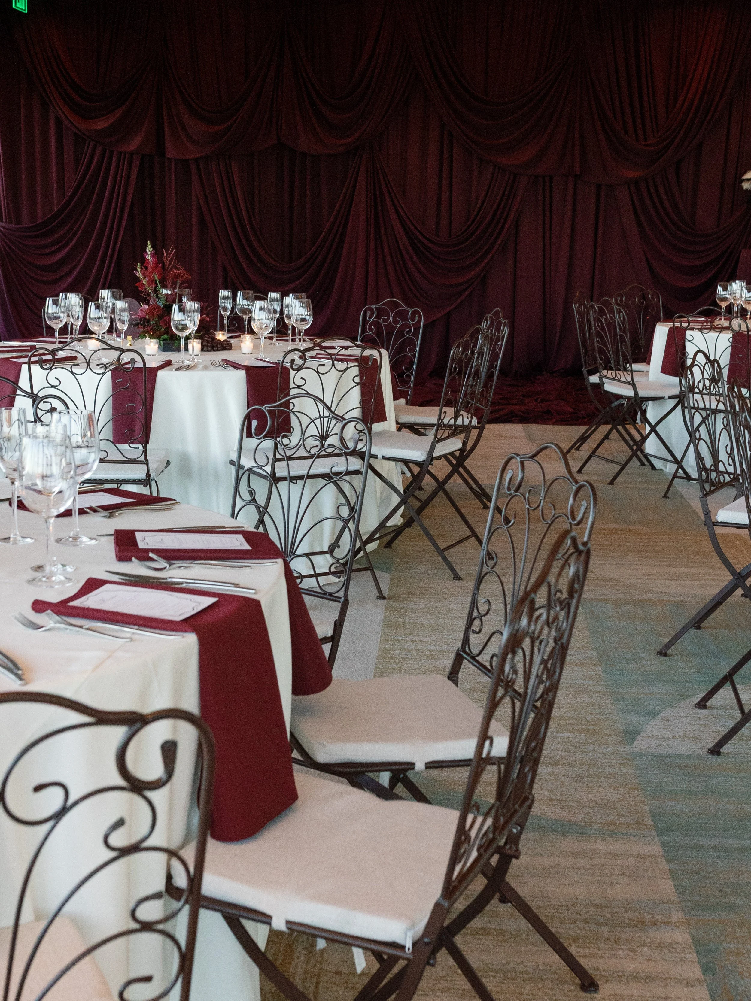 Elegant banquet room with round tables decorated with white tablecloths and burgundy overlays, wine glasses, candles, and floral centerpieces, with a large burgundy curtain backdrop.