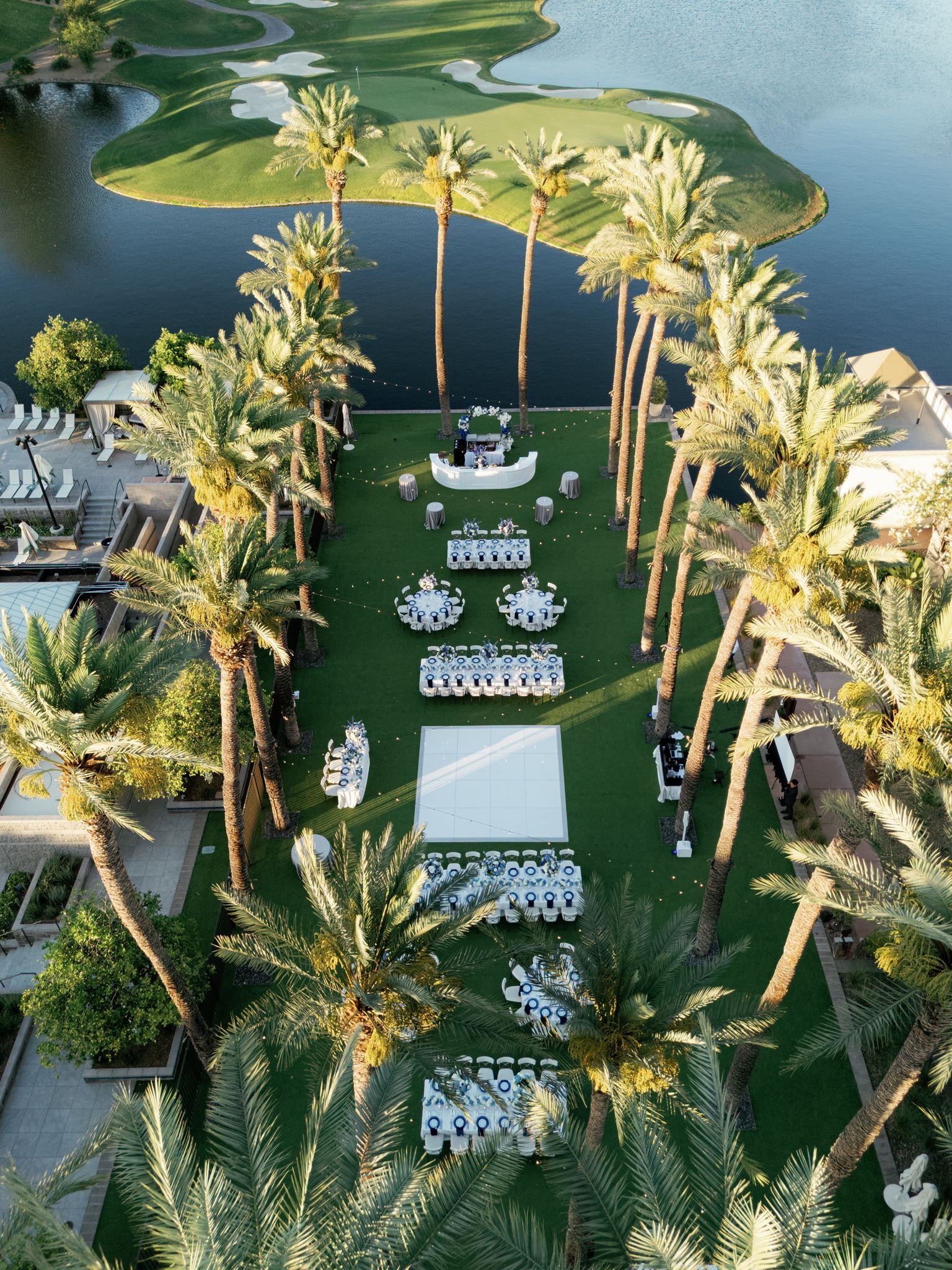 Outdoor event setup with round tables, some decorated with floral arrangements, on a grassy area surrounded by tall palm trees, near a body of water. A white bar and a dance floor are also visible.