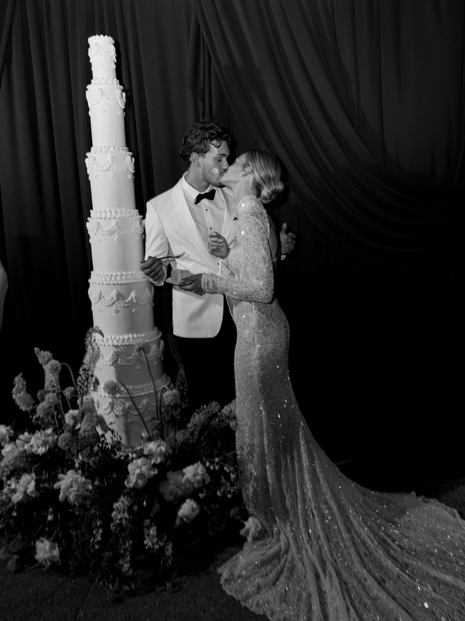 A bride and groom sharing a kiss at their wedding reception, standing beside a tall, decorated wedding cake and a floral arrangement, with dark drapery in the background.