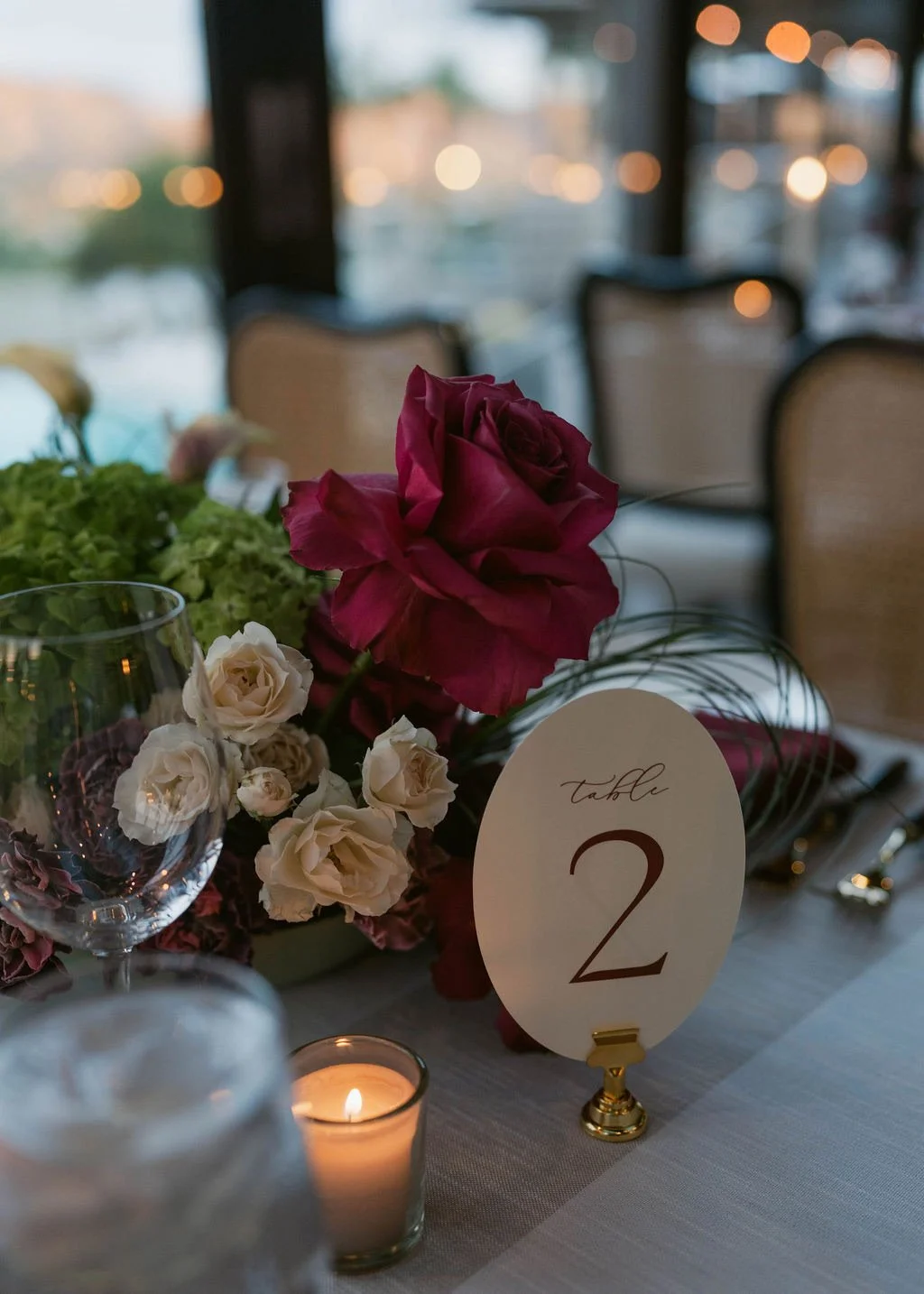 Elegant table setting with a floral centerpiece including a large pink rose and smaller white roses, a candle, wine glasses, and a table number sign indicating table 2, set in a well-lit venue with chairs and large windows in the background.