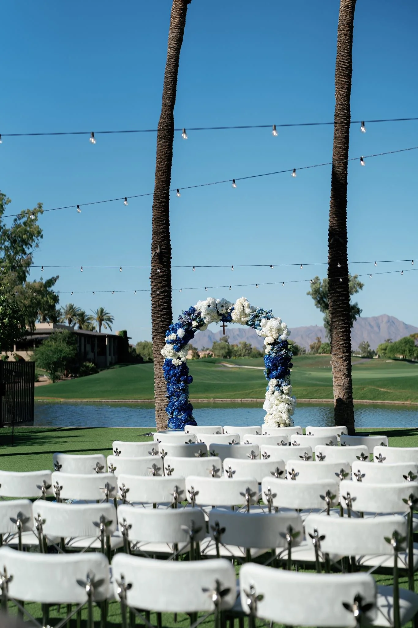 Empty chairs arranged for an outdoor wedding ceremony by a lake with palm trees and mountains in the background. There is an arch decorated with white and blue flowers and a cross, with string lights hanging above.