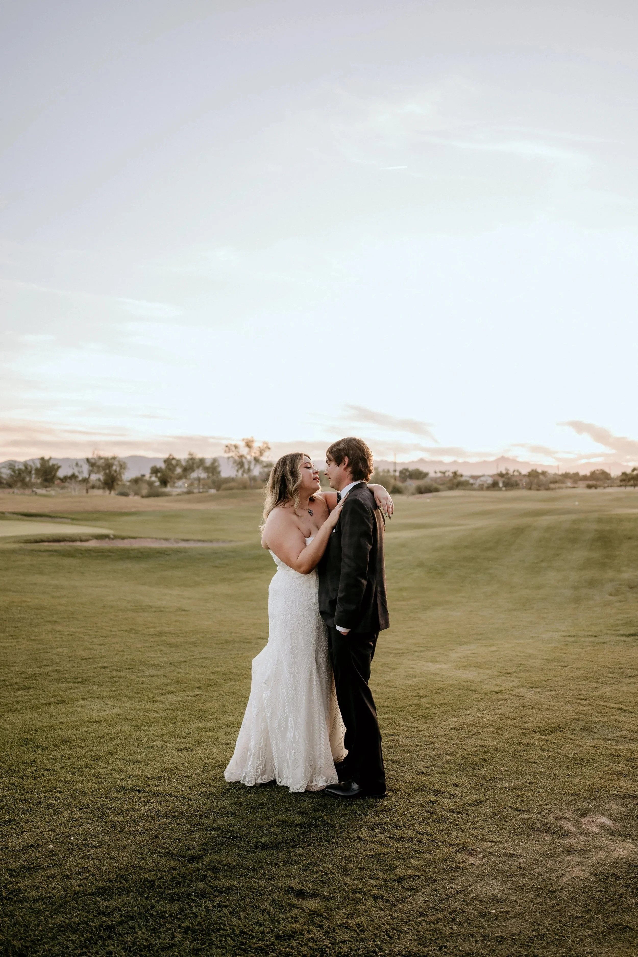 A bride and groom standing on a grassy field during sunset, facing each other closely, with the bride in a white lace wedding gown and the groom in a black suit.