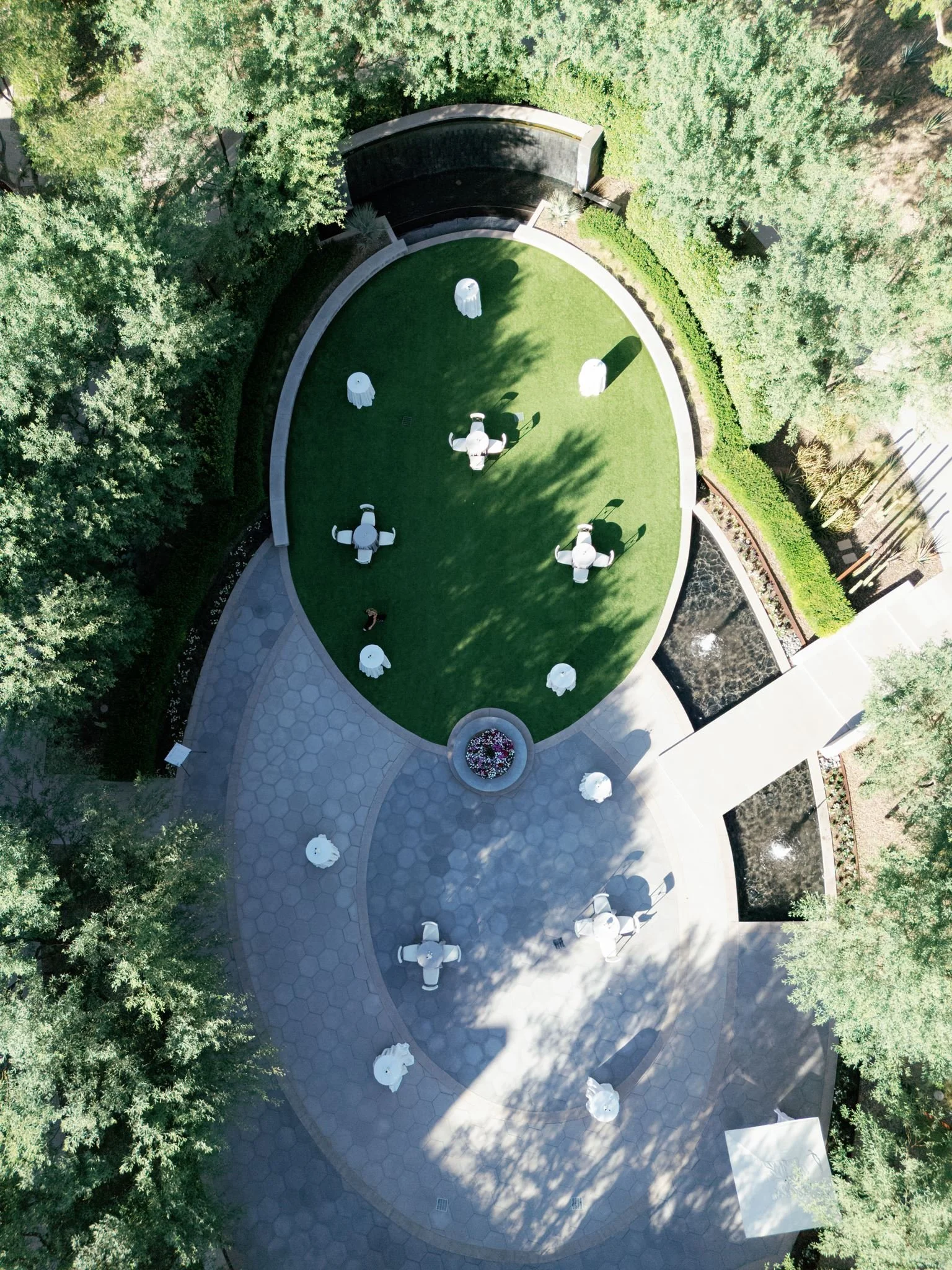 An aerial view of a circular outdoor seating area with white umbrellas around a patch of artificial grass. Surrounding the area are trees and shrubs, with pathways and a small water feature nearby.