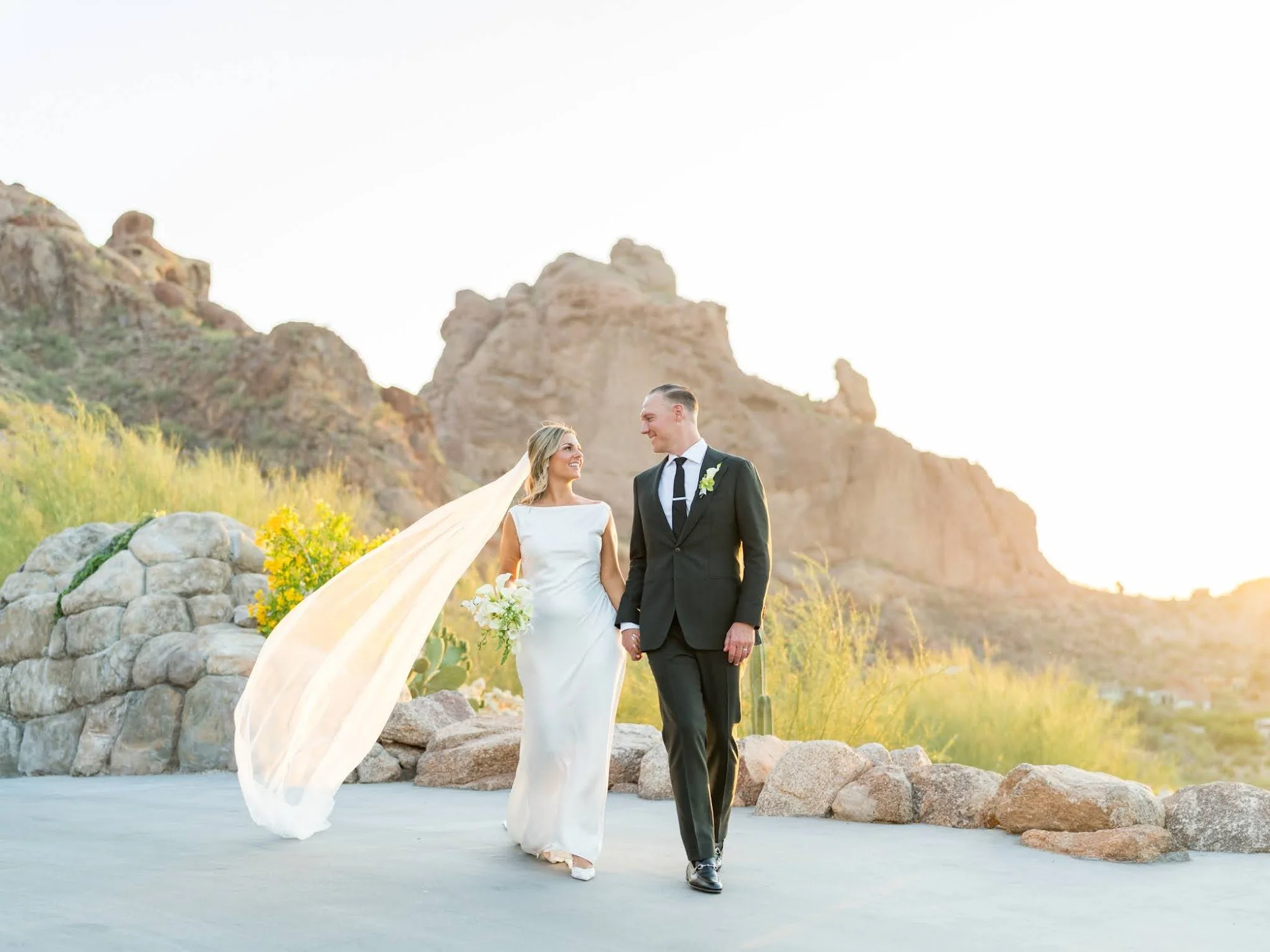 A bride and groom walking hand in hand outdoors during a wedding ceremony at sunset, surrounded by rocks and desert landscape.