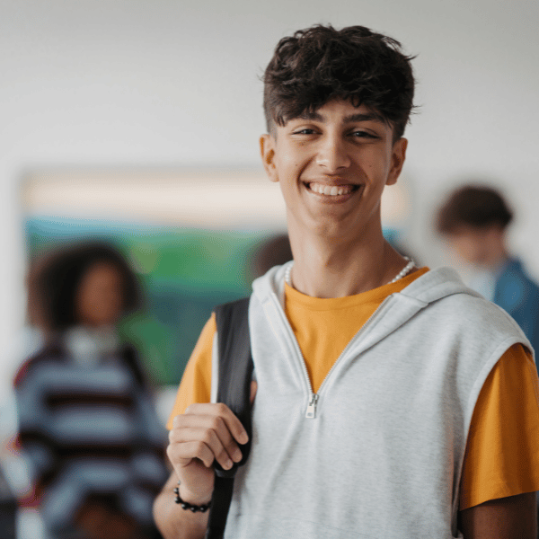 A smiling young man with curly brown hair, wearing a yellow T-shirt and light gray sleeveless hoodie, holding a backpack strap, standing in a school hallway with other students in the background.