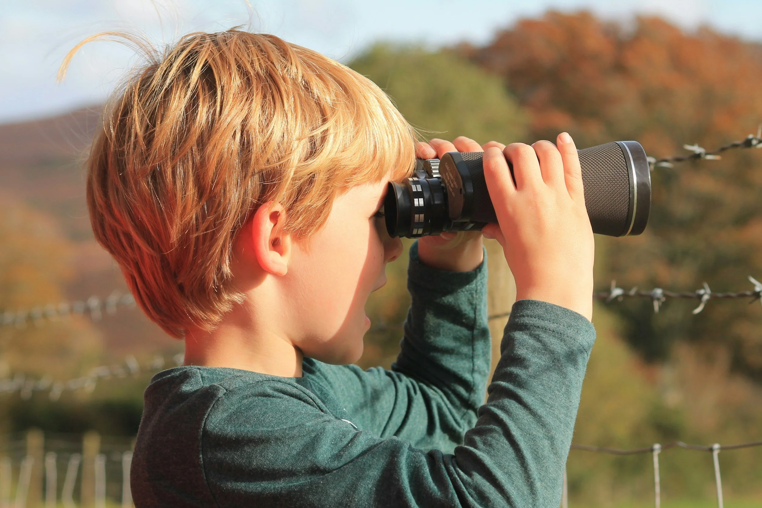Young boy looking through binoculars outdoors with fall foliage in the background.