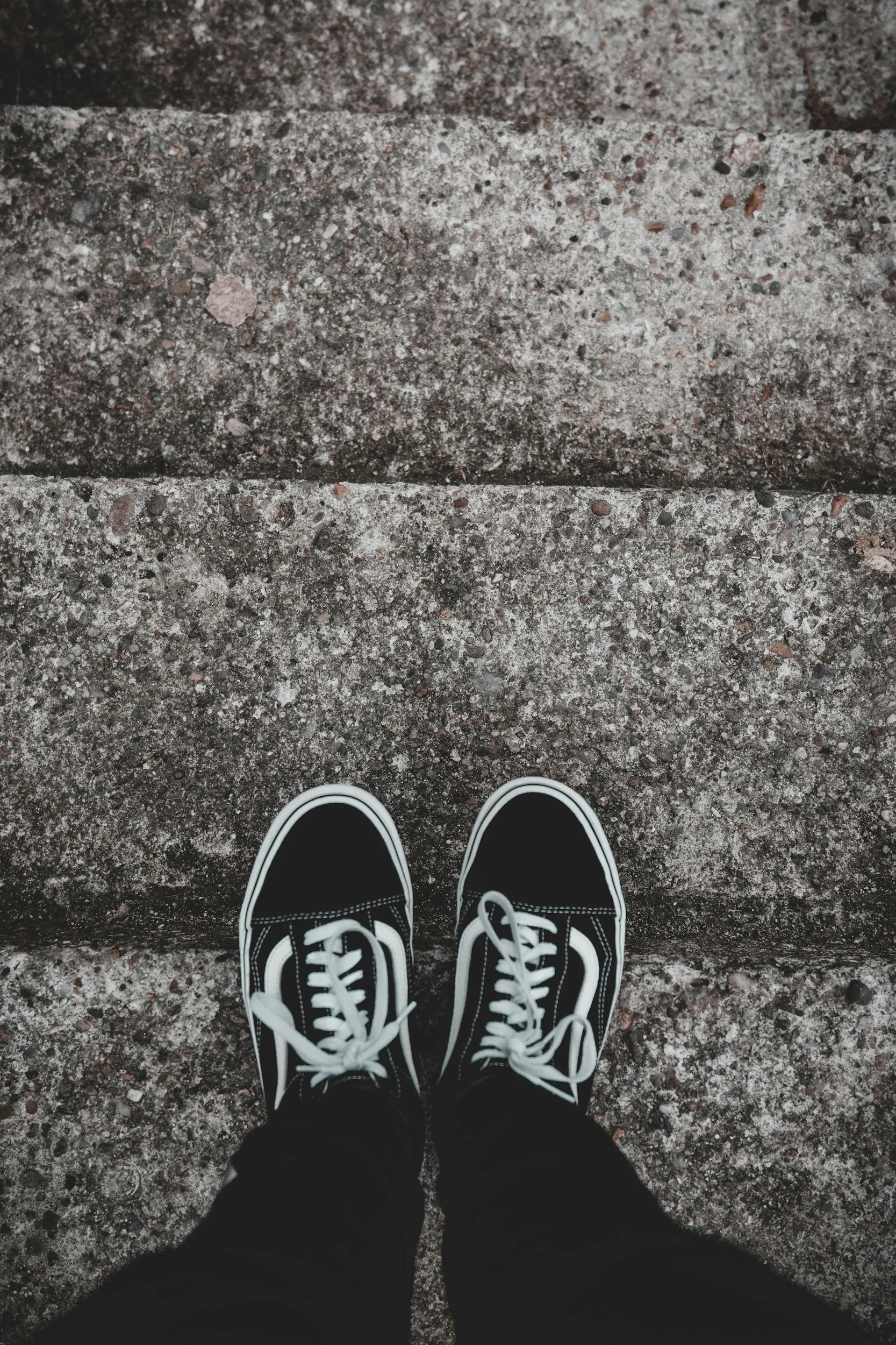 Top view of person's black sneakers with white laces standing on concrete steps.