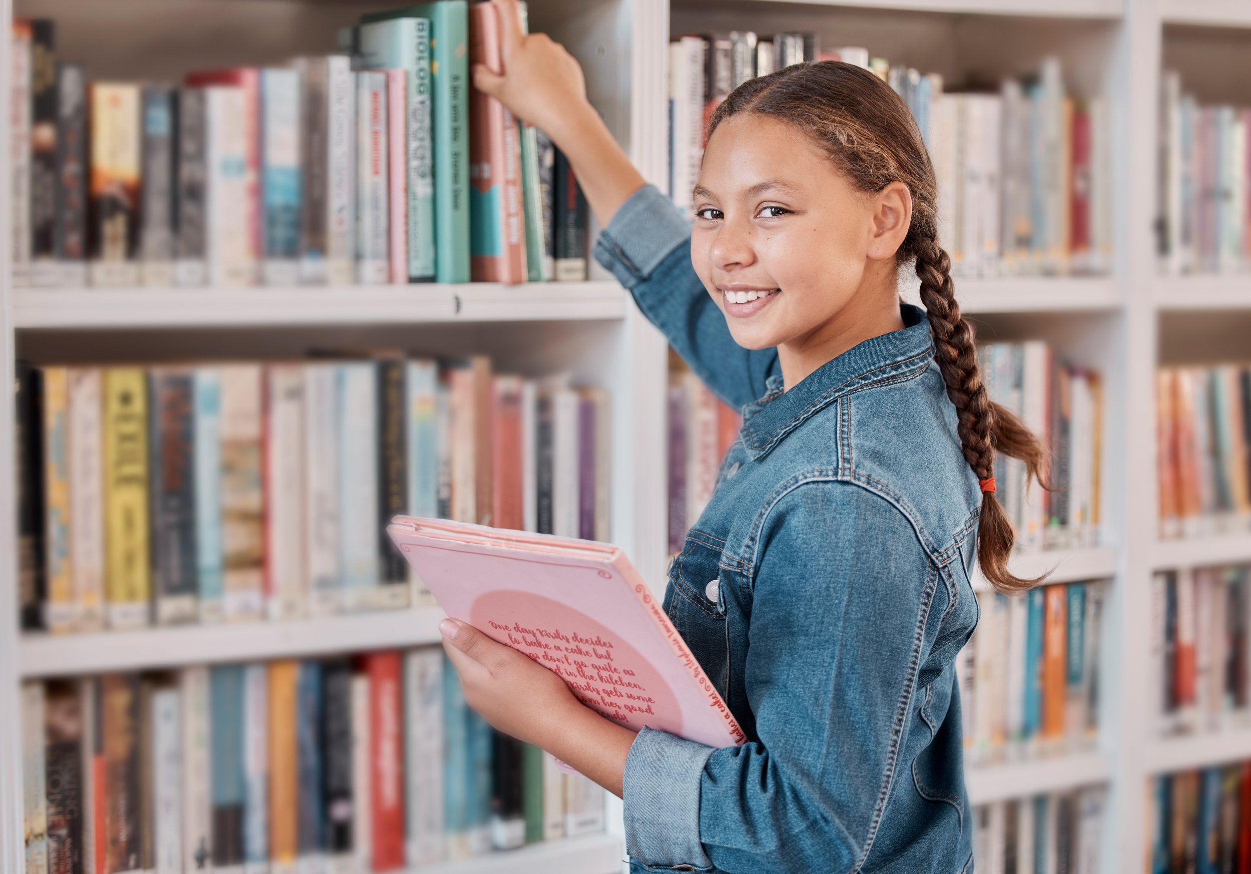 A young girl in a denim jacket with a braid, smiling, holding a pink notebook, reaching for a book on a library shelf.