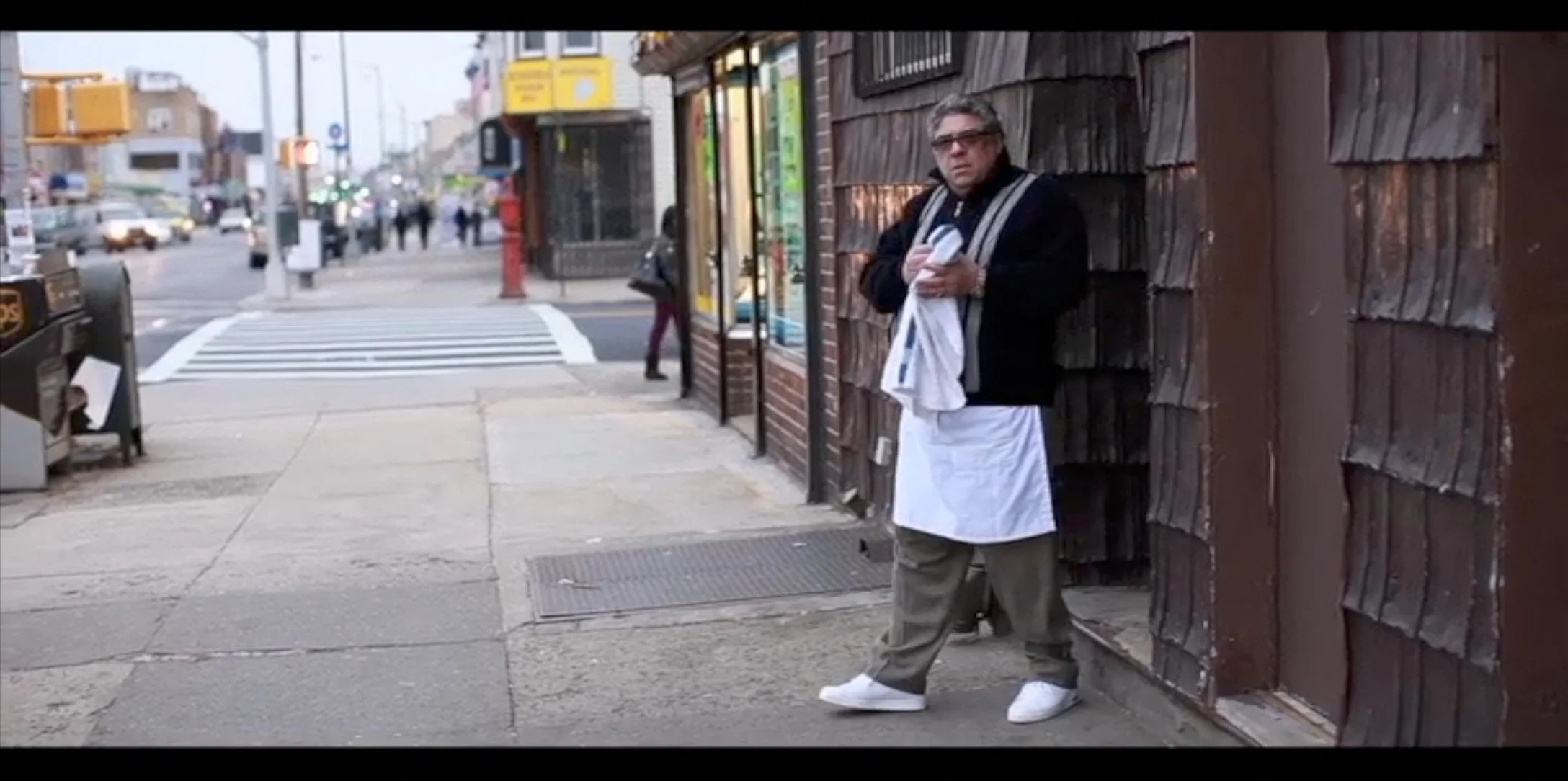 A man wearing glasses and a black jacket with a white apron stands on a city sidewalk, holding some papers or a newspaper, next to a weathered brown wooden wall.
