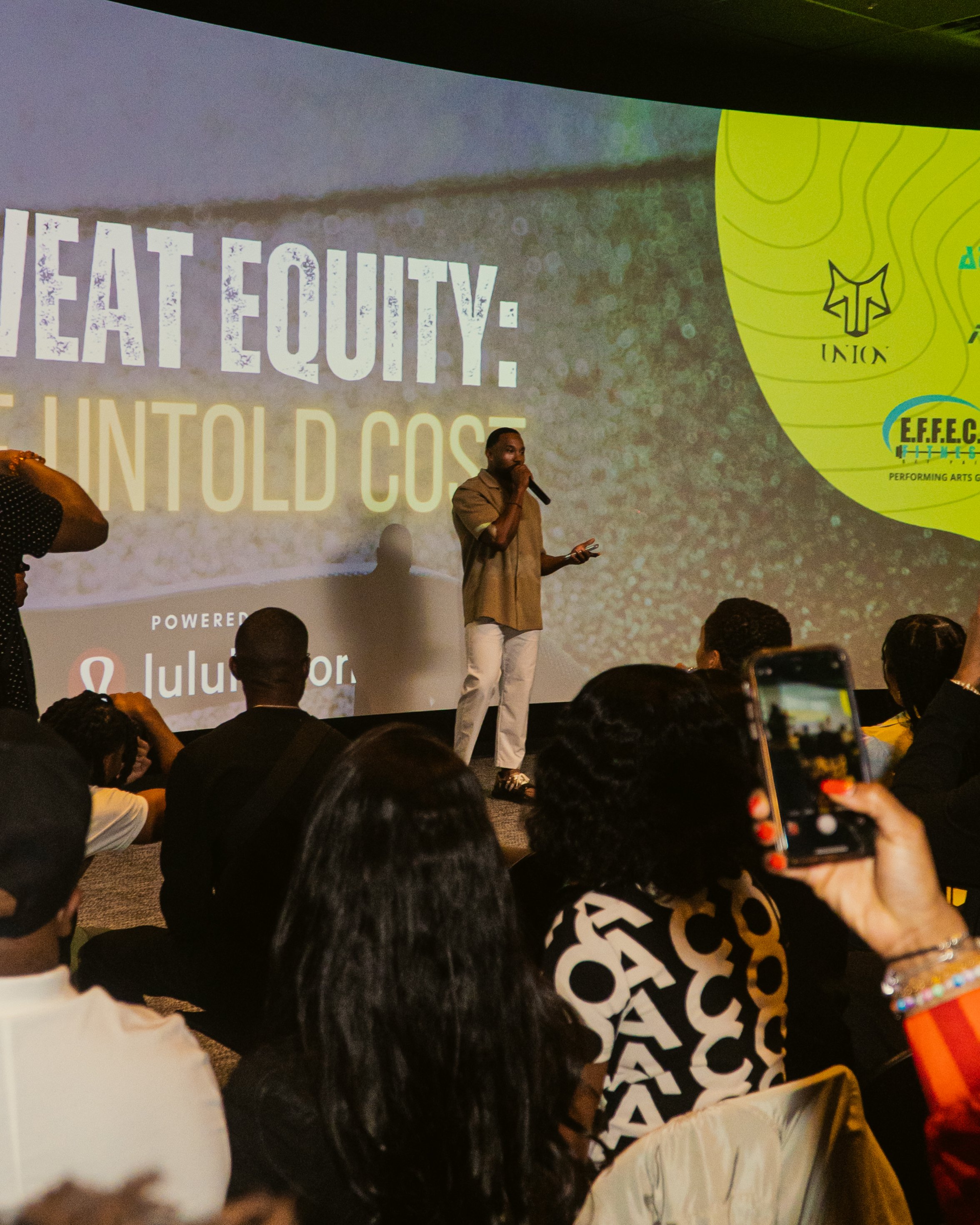 A speaker on stage at a conference or event, standing in front of a large screen displaying the words 'HEALTH EQUITY: UNA TOLD COST' and logos, with an audience seated and taking photos.