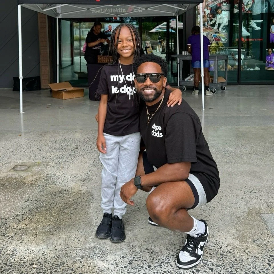 A young girl and a man posing together outside a store under a canopy. The girl has braided hair, is smiling, and is wearing a black t-shirt, light gray sweatpants, and black shoes. The man, squatting and smiling, is wearing sunglasses, a black t-shirt with white text, black shorts, and black and white sneakers.