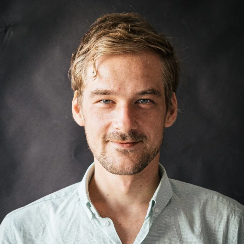 Portrait of a young man with light brown hair, blue eyes, and a beard, wearing a light-colored button-up shirt, standing against a dark background.