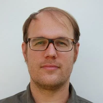Close-up of a man with glasses, light brown hair, and a neutral expression, wearing a gray shirt against a plain light background.