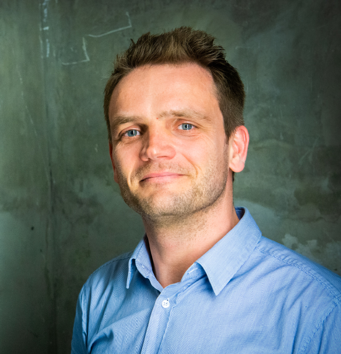 A portrait of a man wearing a light blue button-up shirt, standing against a textured green wall.