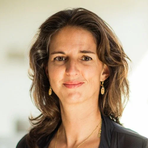 Headshot of a woman with shoulder-length wavy brown hair, wearing gold earrings and a black top, smiling softly at the camera.