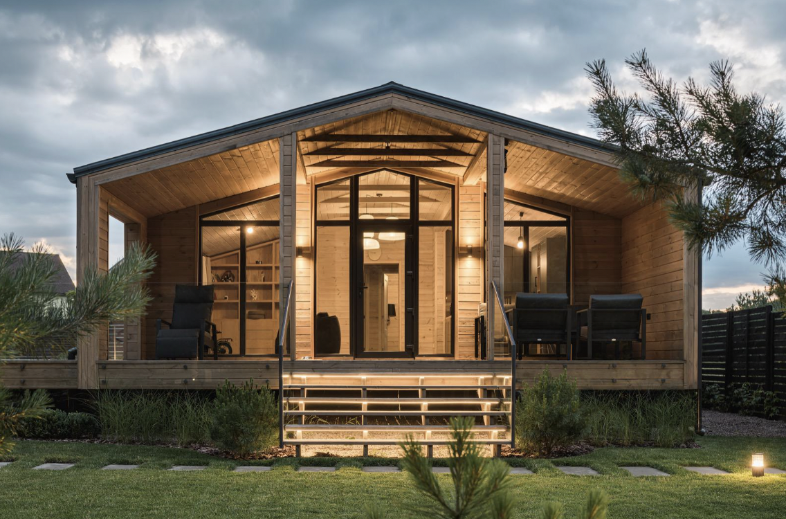 A modern wooden house with an illuminated front porch, large glass doors and windows, surrounded by greenery at dusk.