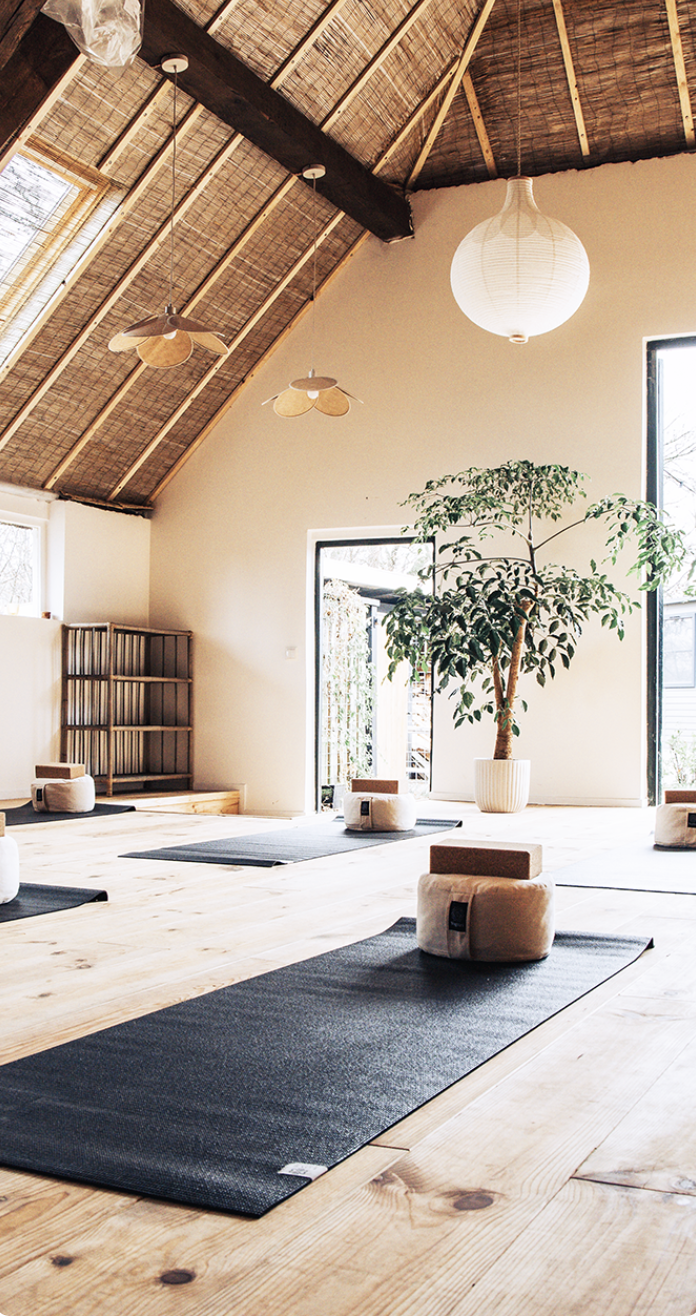 A yoga or meditation studio with wooden floors, black mats, and wrapped yoga blocks. Large potted plant near open doors, with pendant and wall-mounted light fixtures on a high ceiling with exposed wooden beams.