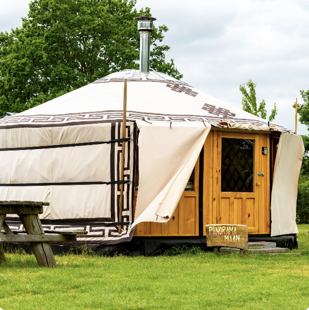 A yurt with a wooden door and a sign that reads 'Panorama Maan,'set on green grass with a picnic table nearby, trees and cloudy sky in the background.