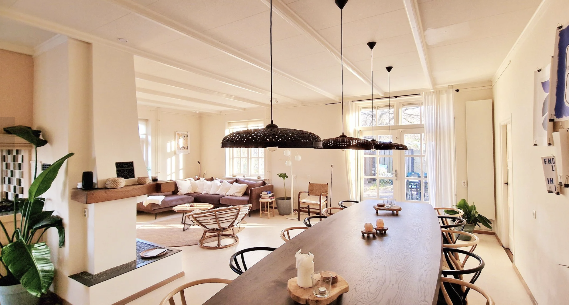Bright open living room and dining area with large windows, beige walls, white ceiling, and wooden furniture. A long wooden dining table with black chairs, pendant lights above, and a cozy seating area with a sectional sofa, chairs, and plants.