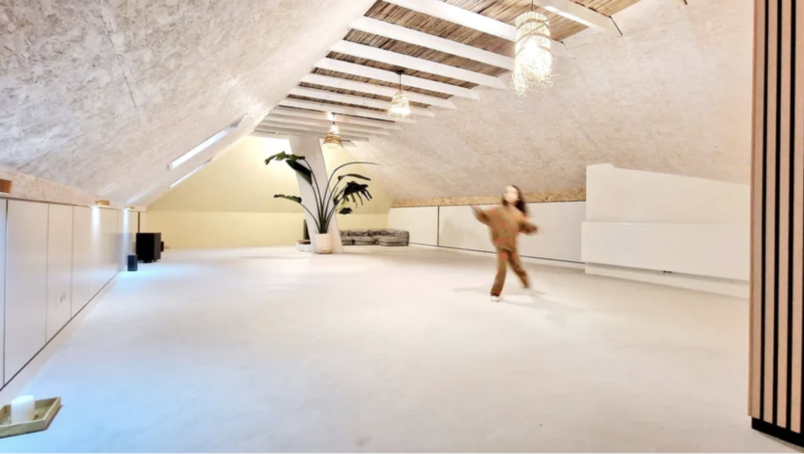Child playing in a spacious, modern attic room with sloped ceiling, wooden beams, hanging pendant lights, large plant, and minimal decor.