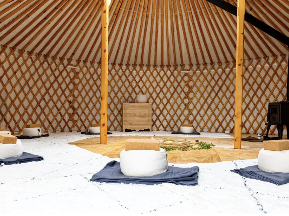 Inside a yurt with wooden lattice walls and ceiling, decorated with cushions and a small wooden table, set up for a meditation or yoga session.