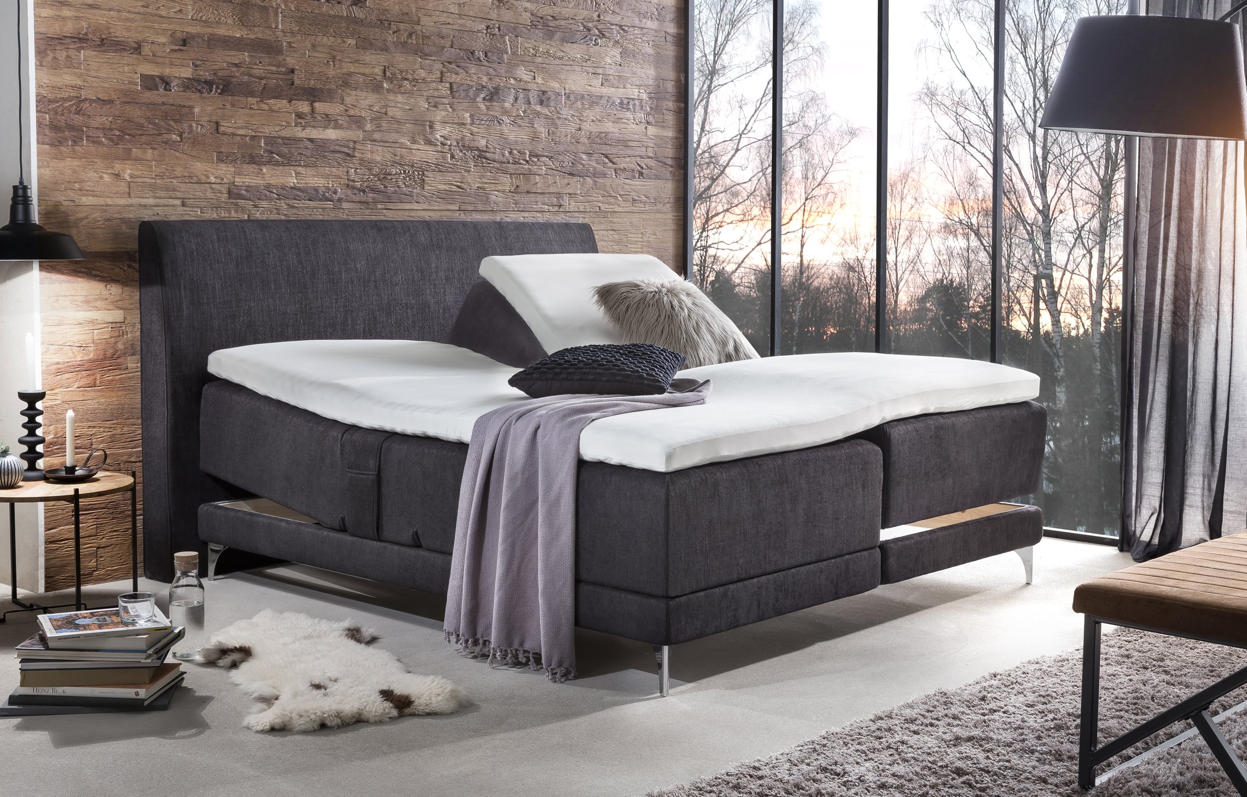 Modernes Schlafzimmer mit Bett, großen Fenstern, Stadtblick, Fahrrad, Pflanze, Bücher auf Boden, graue Akzentwand, Holztisch mit Dekoration.