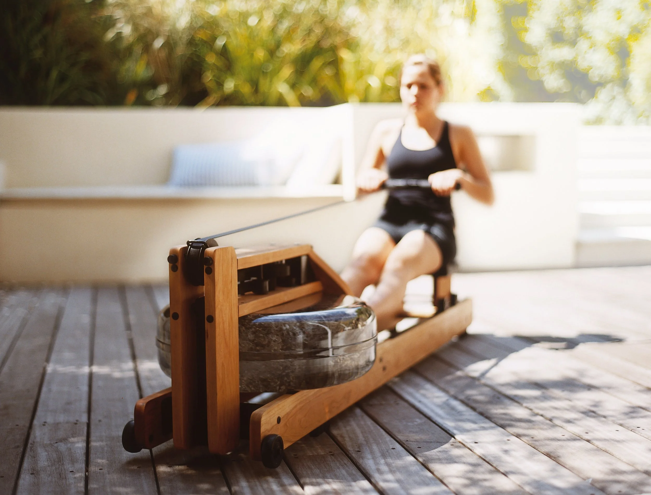 Person benutzt ein Rudergerät in einem hellen Raum mit Holzboden und Pflanzen im Hintergrund.