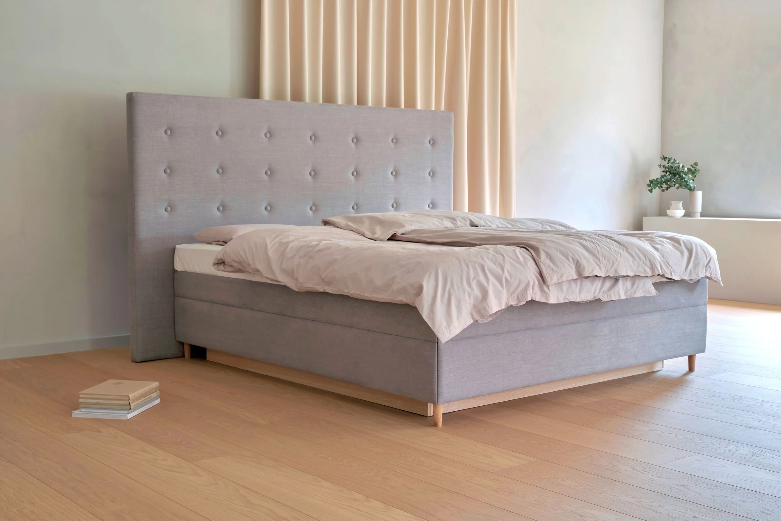 Minimalistisches Schlafzimmer mit Bett, grauer Boxspring-Matratze, beigem Bettlaken und Kopfkissen mit leichter Unordnung. Im Hintergrund ein heller Vorhang und ein paar Dekorationspflanzen auf einem Schrank.