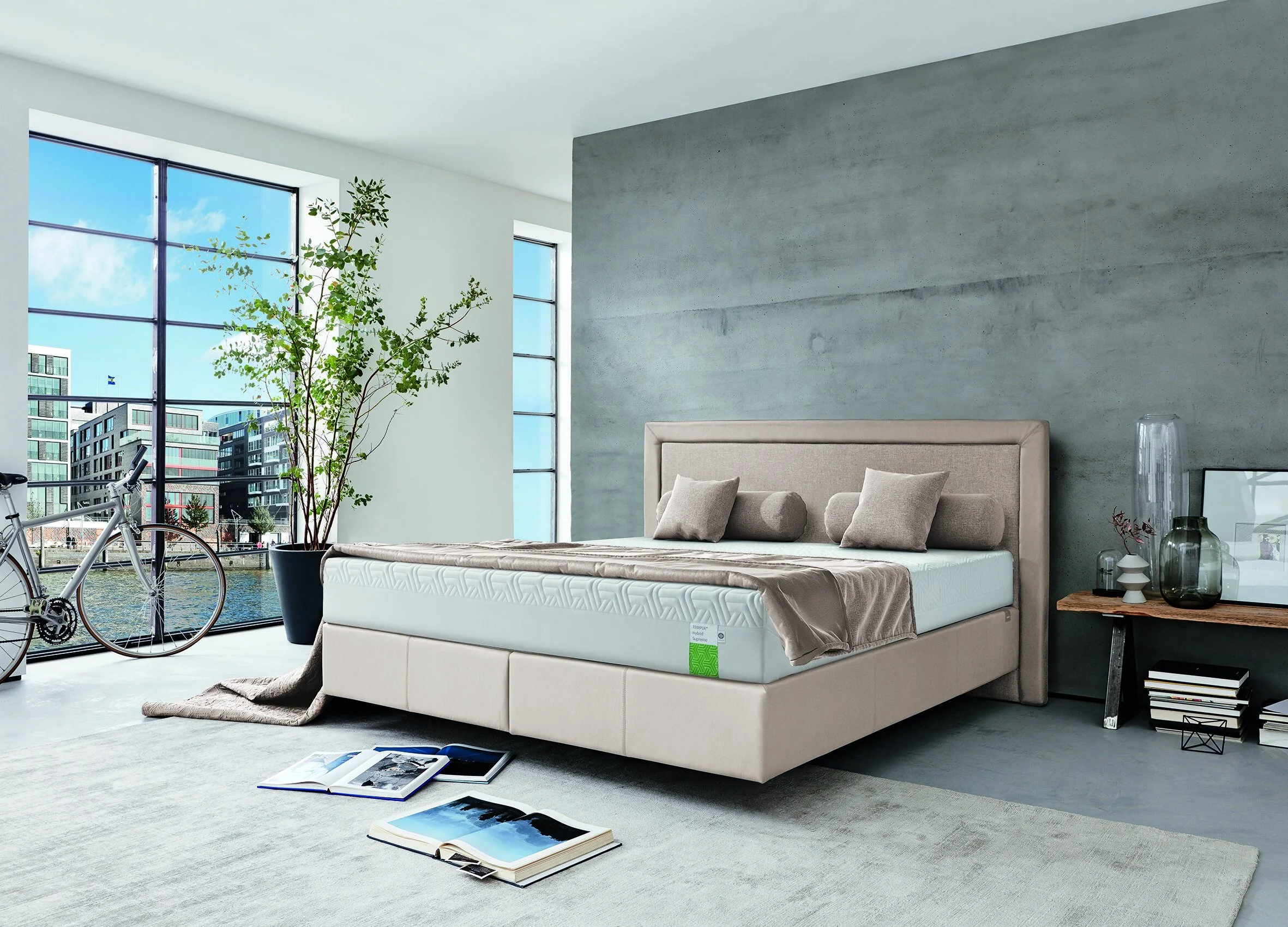 Modernes Schlafzimmer mit Bett, großen Fenstern, Stadtblick, Fahrrad, Pflanze, Bücher auf Boden, graue Akzentwand, Holztisch mit Dekoration.