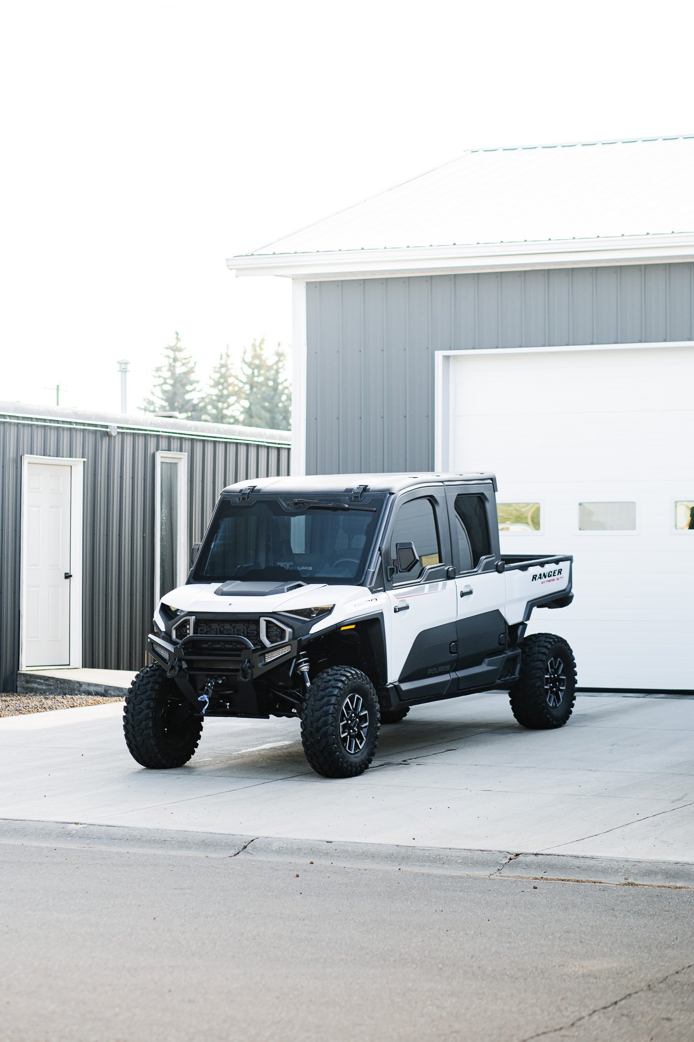 A white and black Polaris Ranger off-road vehicle with tinted windows parked outside a gray building with a white door and garage door in Hanna, Alberta.