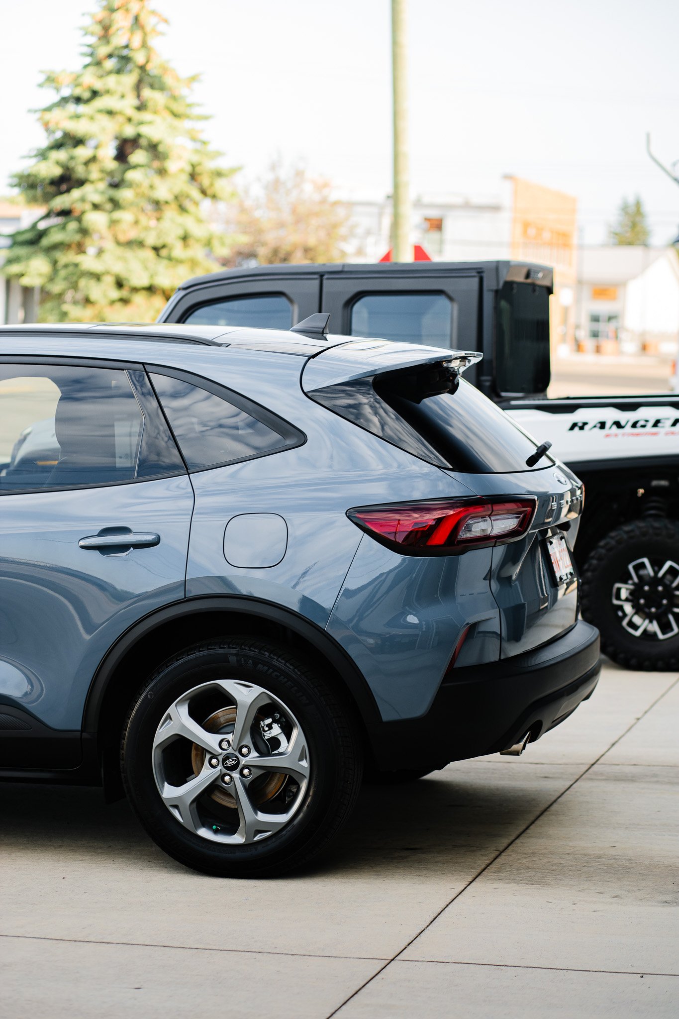 Close-up of a modern blue Ford Escape SUV with paint protection film and ceramic coating parked on a concrete driveway, with a gray pickup truck behind it, and trees and buildings in the background in Hanna, Alberta.