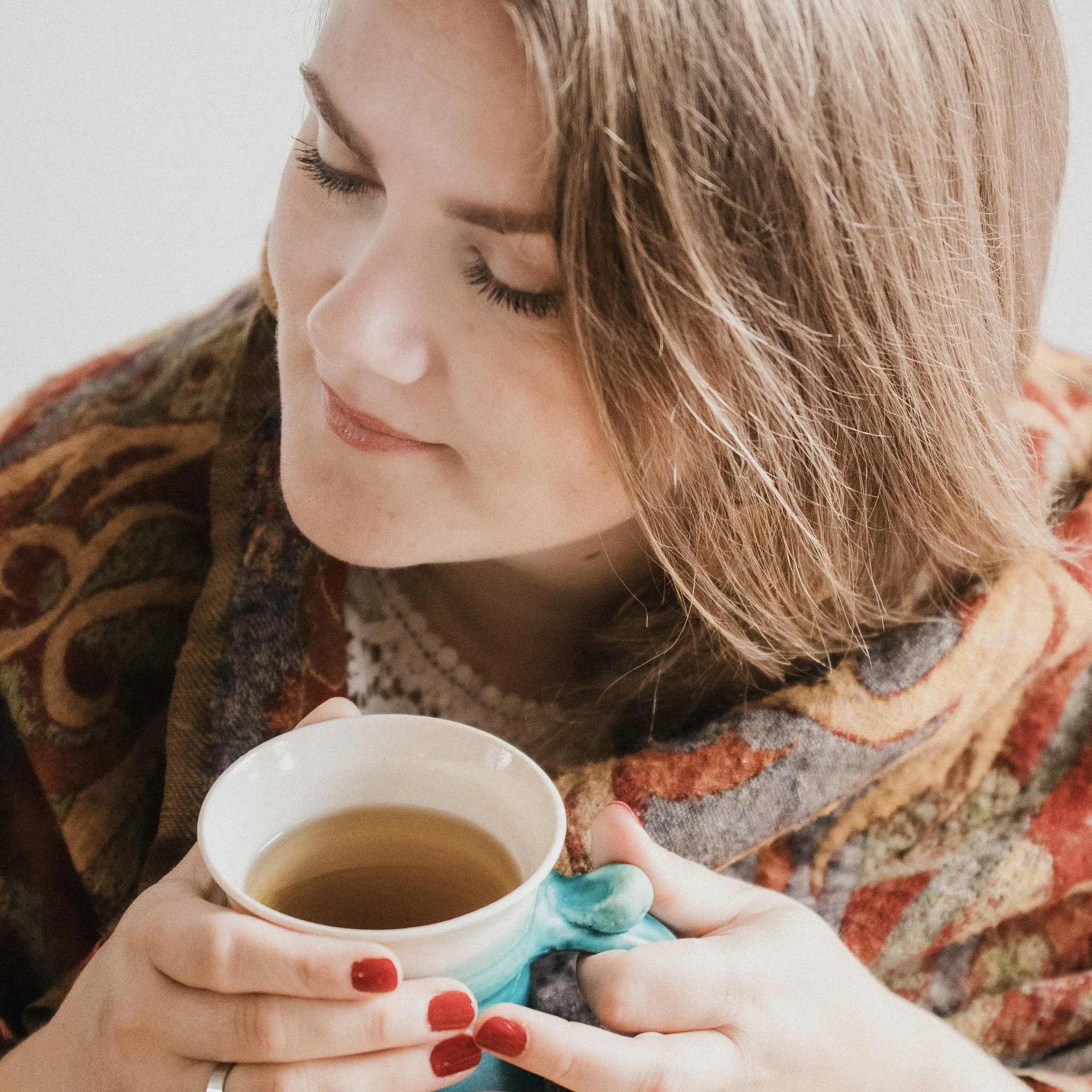 Person with red nail polish holding a white mug of hot coffee or tea while standing on a patterned rug.