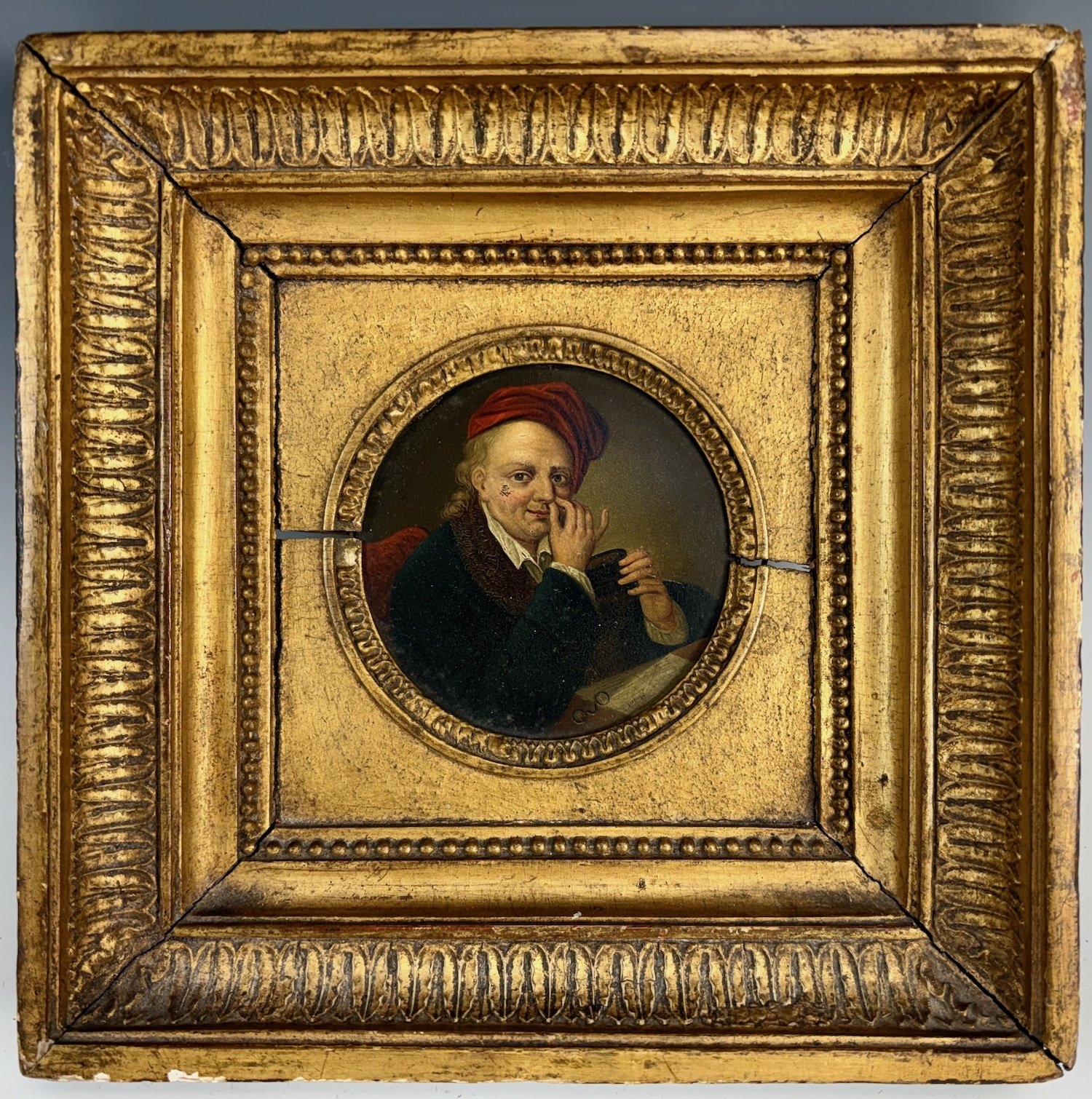 A small, circular portrait of a man with curly hair, wearing a red cap and dark clothing, holding a book and touching his nose with his finger. The portrait is surrounded by an ornate, gold, square frame with intricate detailing, including bead and leaf patterns.