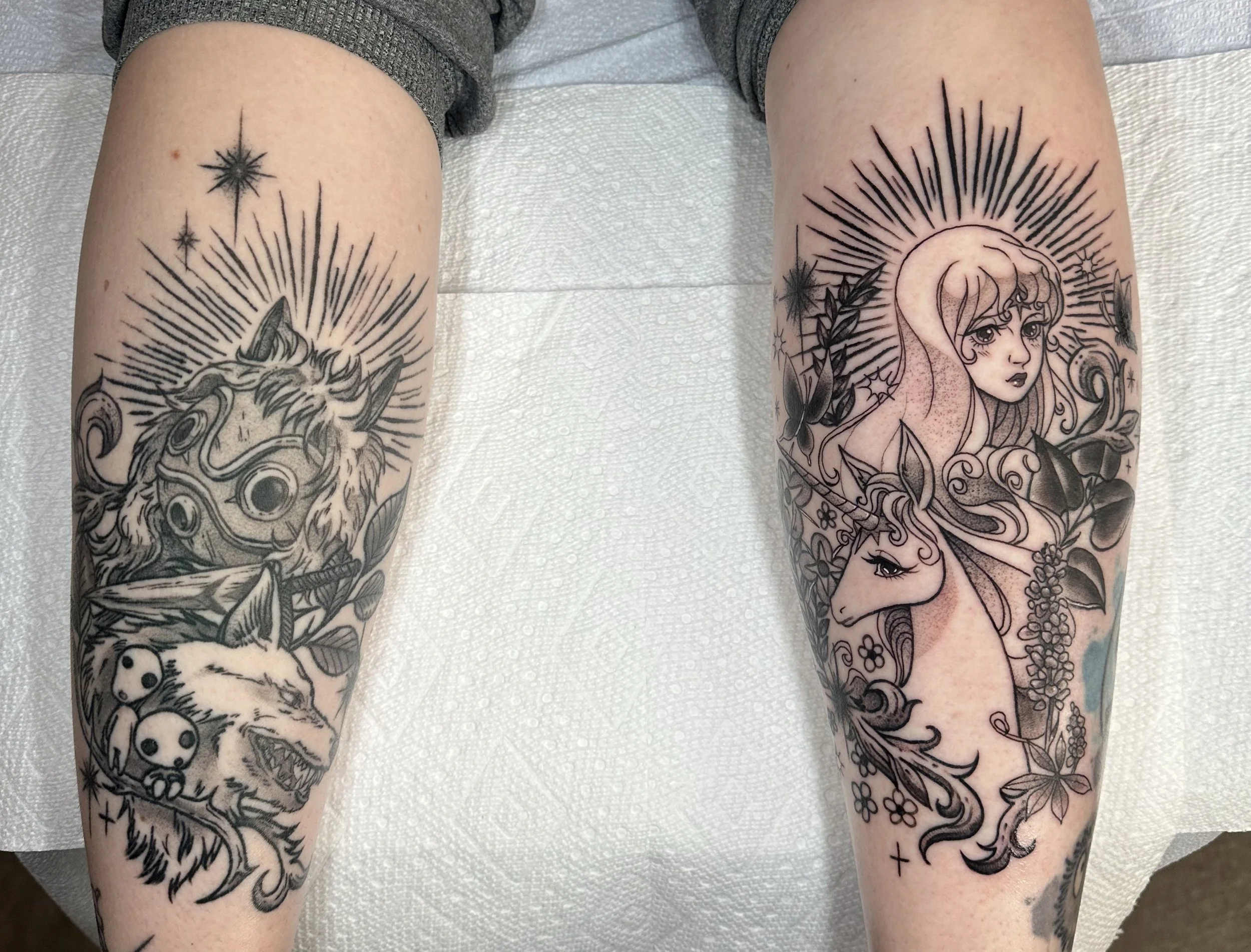 Black and grey tattoos on the back of the calfs, one is Princess Mononoke with Moro, and kodama forest spirits, the other is the Last Unicorn with plants and Lady Amalthea  by Portland Tattoo artist at Upper Moon Tattoo Rae Syndulla 