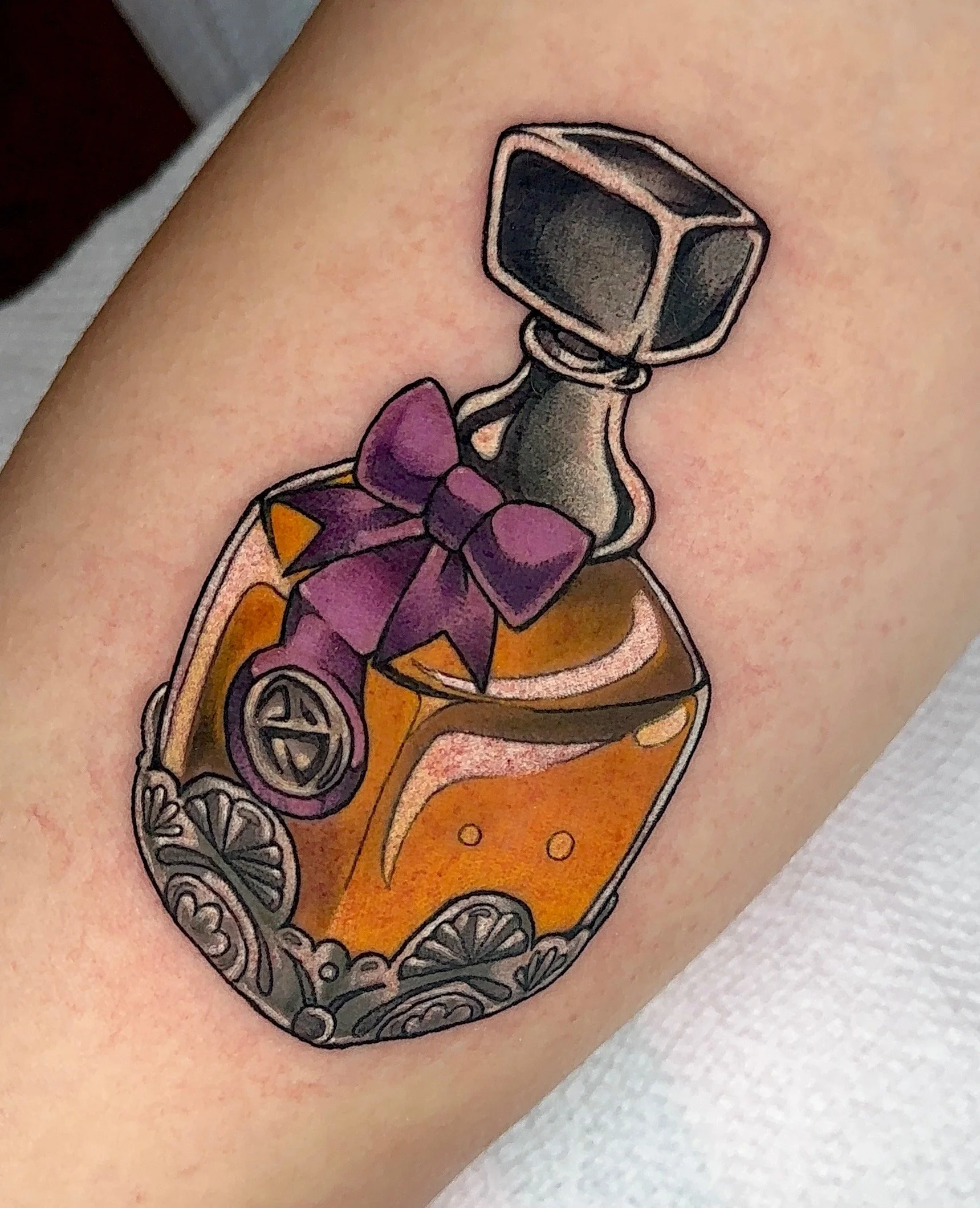 Color tattoo of a fancy bottle with a bow and metal embellishments on the base, liquor bottle, potion bottle  by Portland Tattoo artist at Upper Moon Tattoo Rae Syndulla 