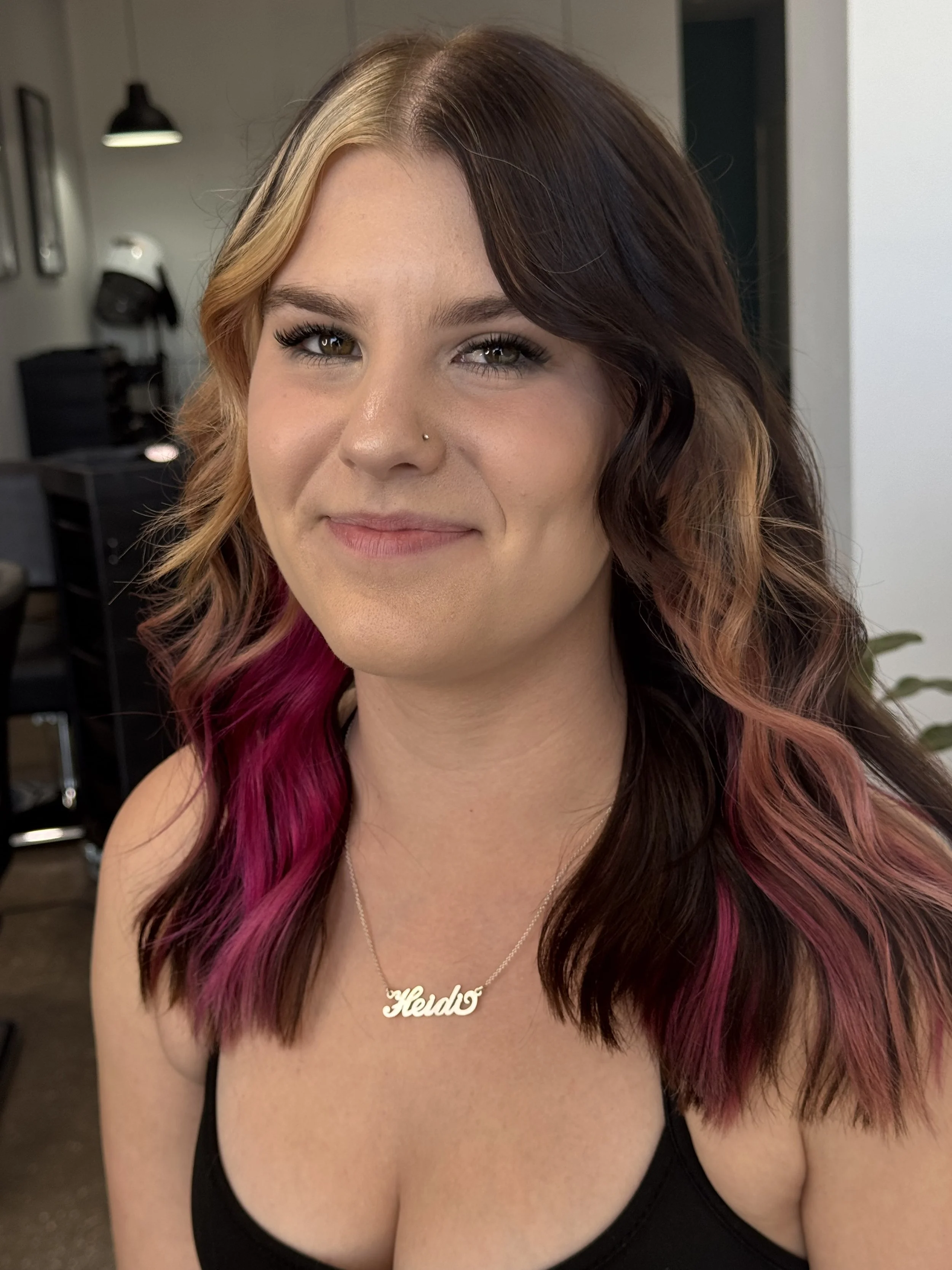 A young woman with wavy, brown hair with pink highlights, wearing a necklace that says 'Hedo', smiling at the camera in a salon or studio setting.