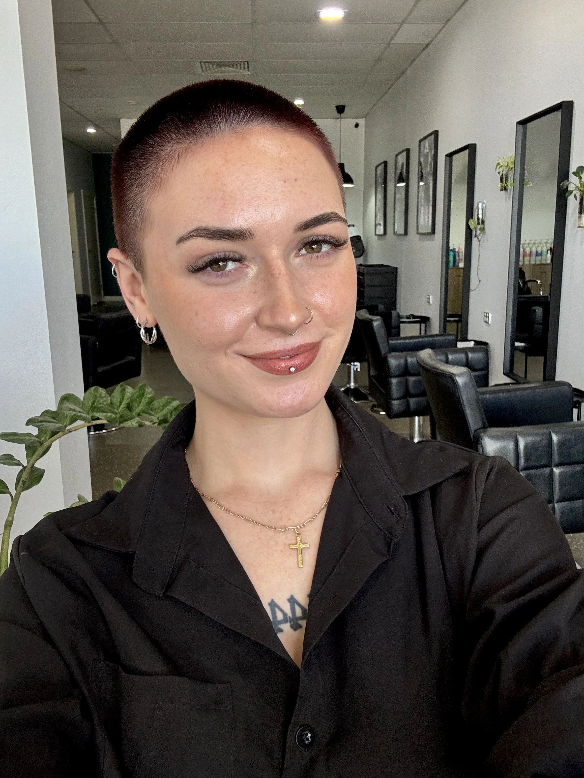 A woman with short buzzed hair, piercings, and tattoos, smiling in a hair salon with black chairs and mirrors in the background.