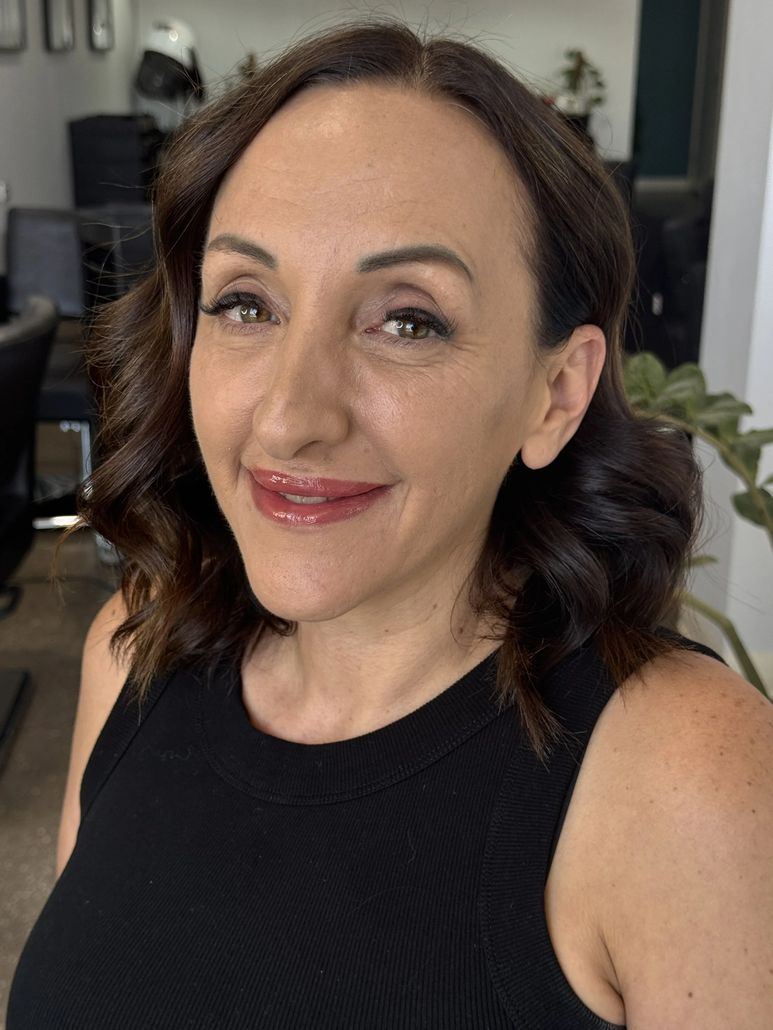 A woman with shoulder-length wavy brown hair and light skin, smiling in an indoor setting, wearing a black sleeveless top.