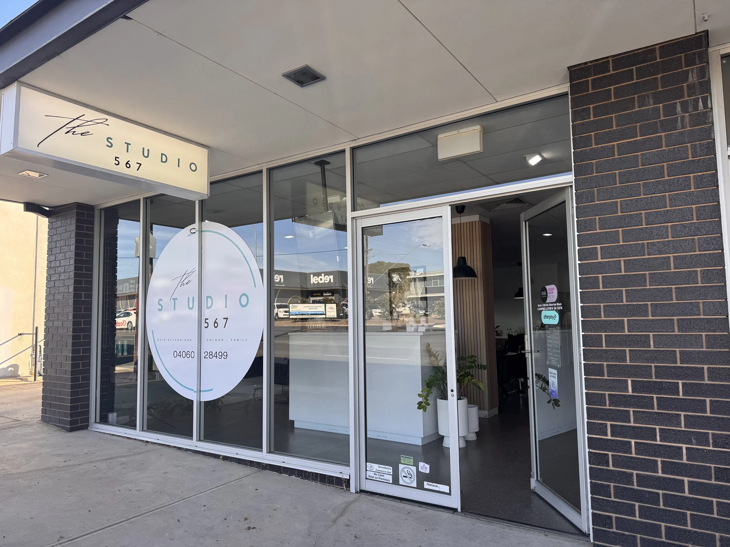 Front view of a hair salon called 'The Studio 567', with glass windows, a hanging sign, and a door open to the interior. Potted plants are visible inside.