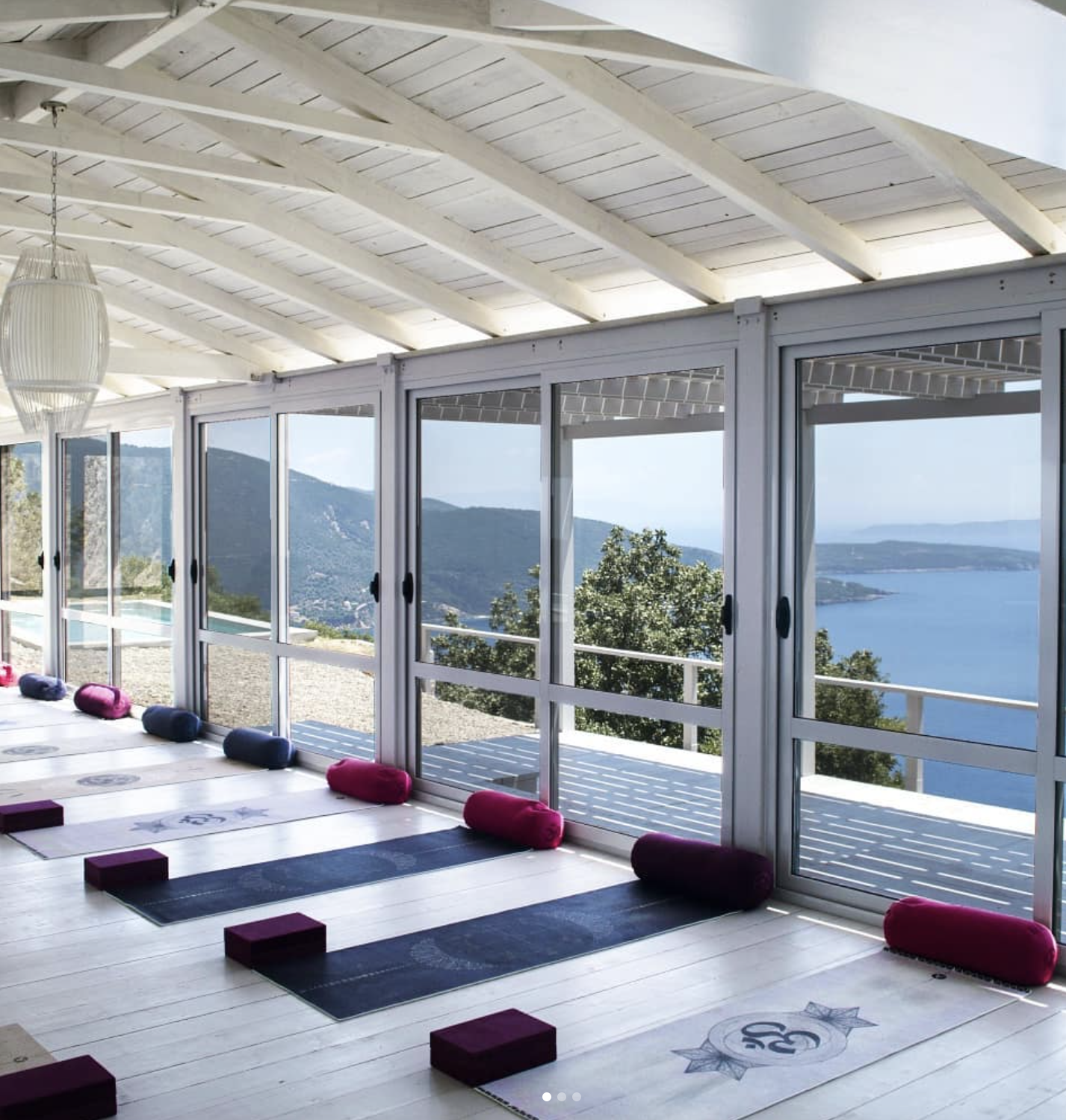 Yoga studio with mats, bolsters, and blocks, overlooking a scenic lake and mountains through large glass windows.