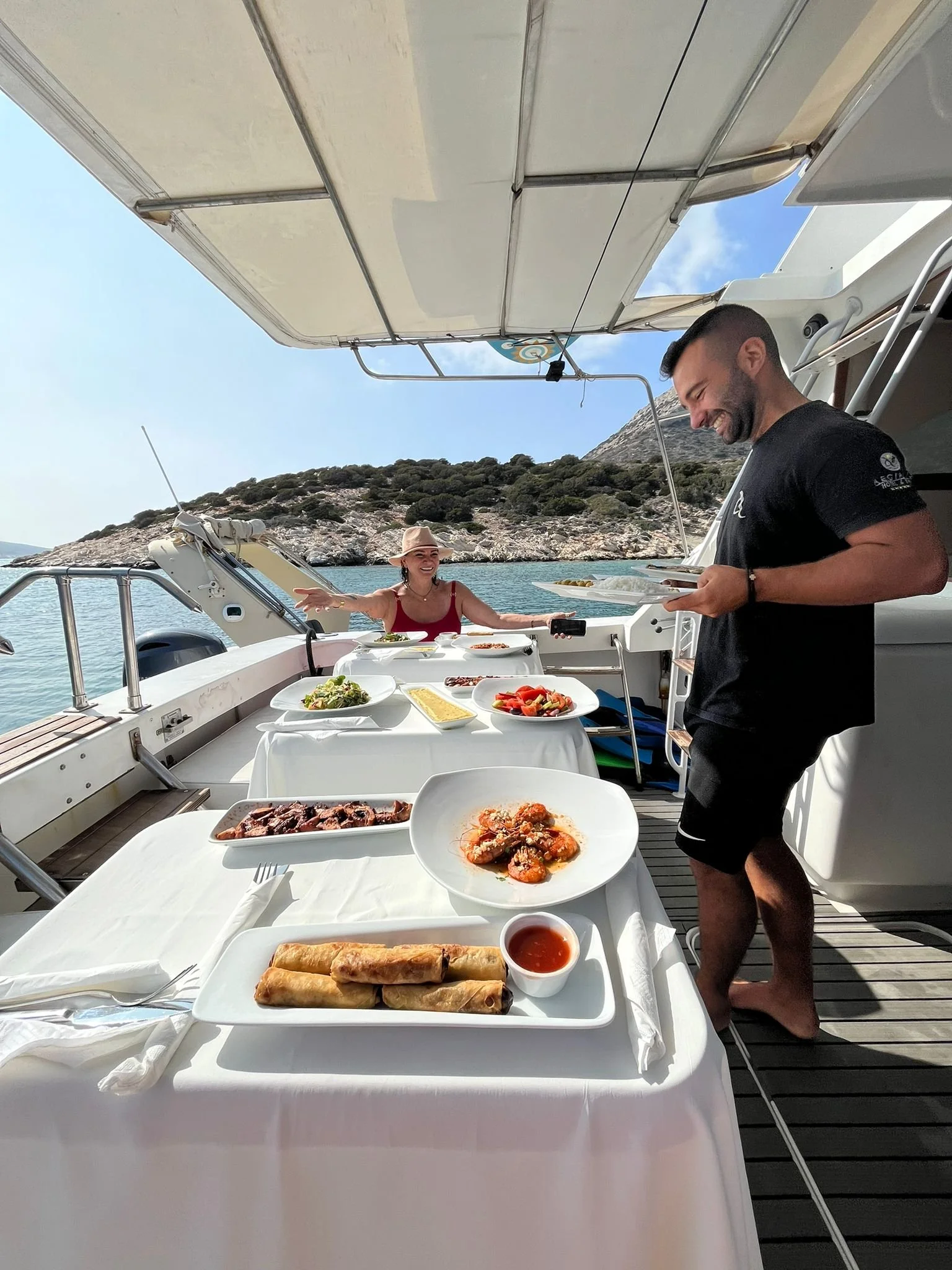 People enjoying a meal on a yacht with scenic ocean and rocky hillside in the background.