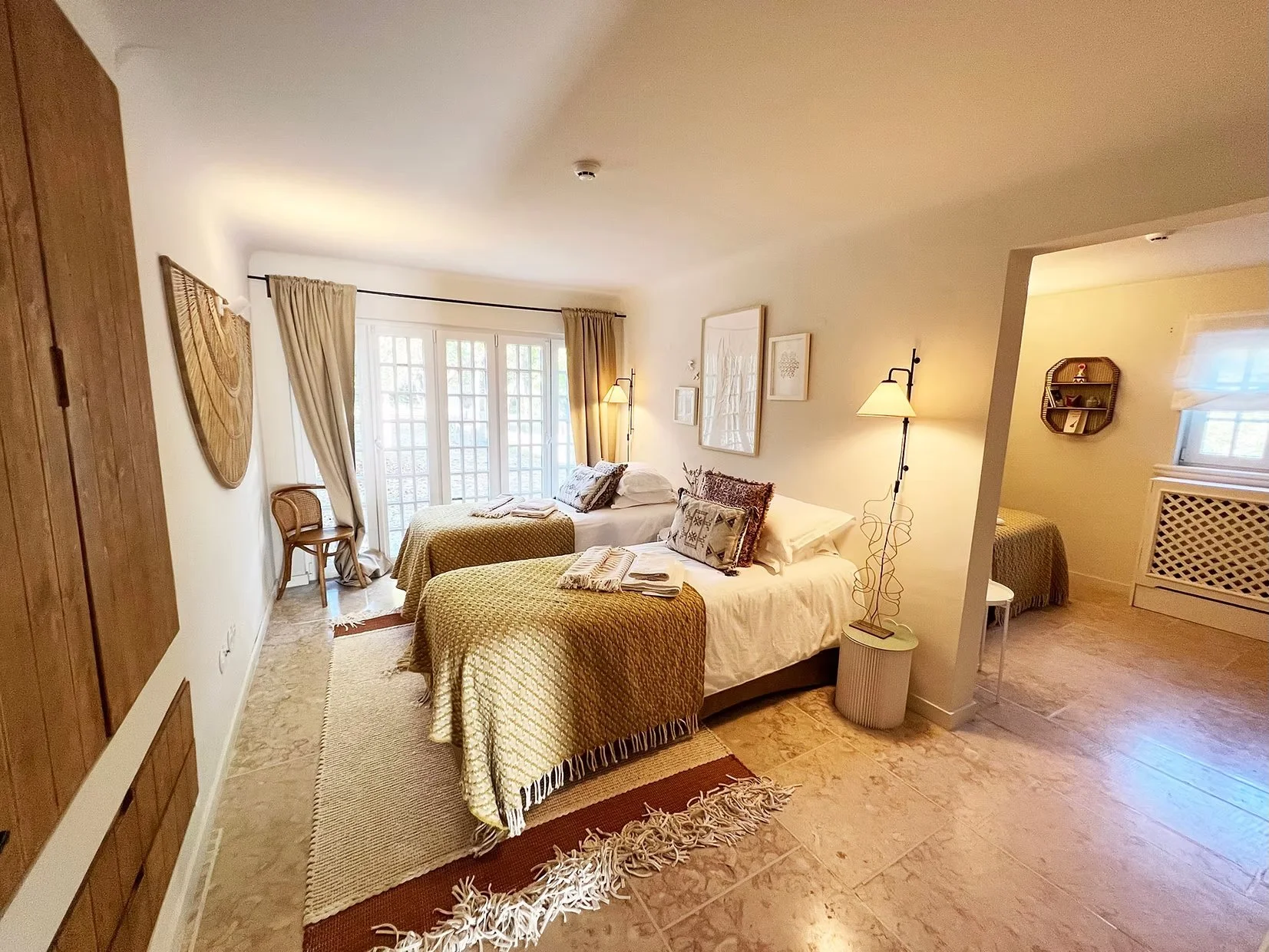 A cozy bedroom with two single beds, each covered with a gold-colored quilt, and white sheets and pillows. There are towels on each bed. The room has a large window with glass doors, beige curtains, and a small chair nearby. Artistic wall decorations and two wall-mounted lamps provide lighting. A small side table with a lamp is beside the bed. There is an adjoining smaller room with a window and a small wall shelf. The flooring consists of beige tiles, and there is a beige rug on the floor.