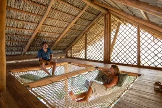 Two women relaxing on a hammock inside a wooden hut with a thatched roof and lattice walls.