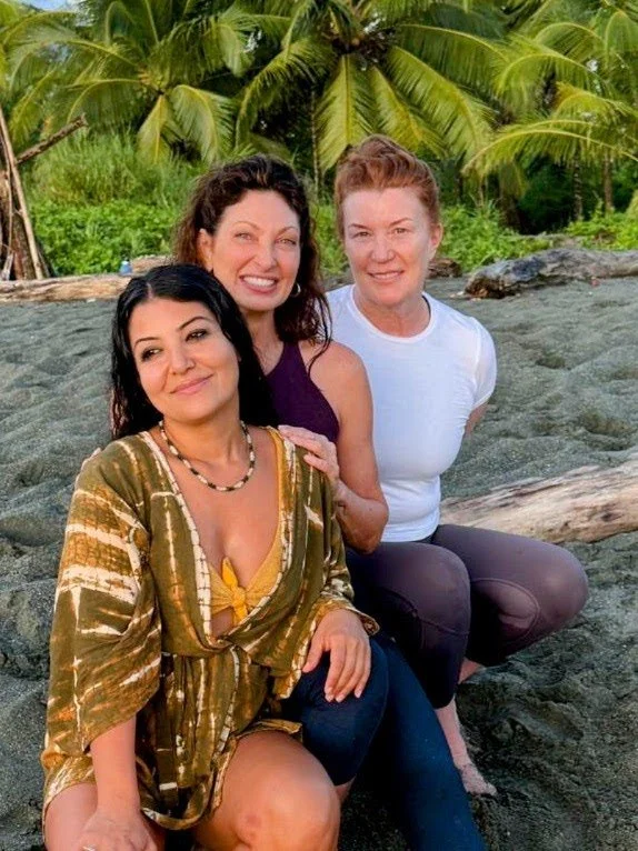Three women sitting on a rocky beach with tropical palm trees in the background.