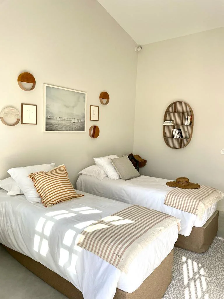 A bedroom with two twin beds dressed in white bedding, striped pillows, and matching striped throws. There is wall art above the beds and round wooden shelves with books on the right side. A straw hat rests on one bed.