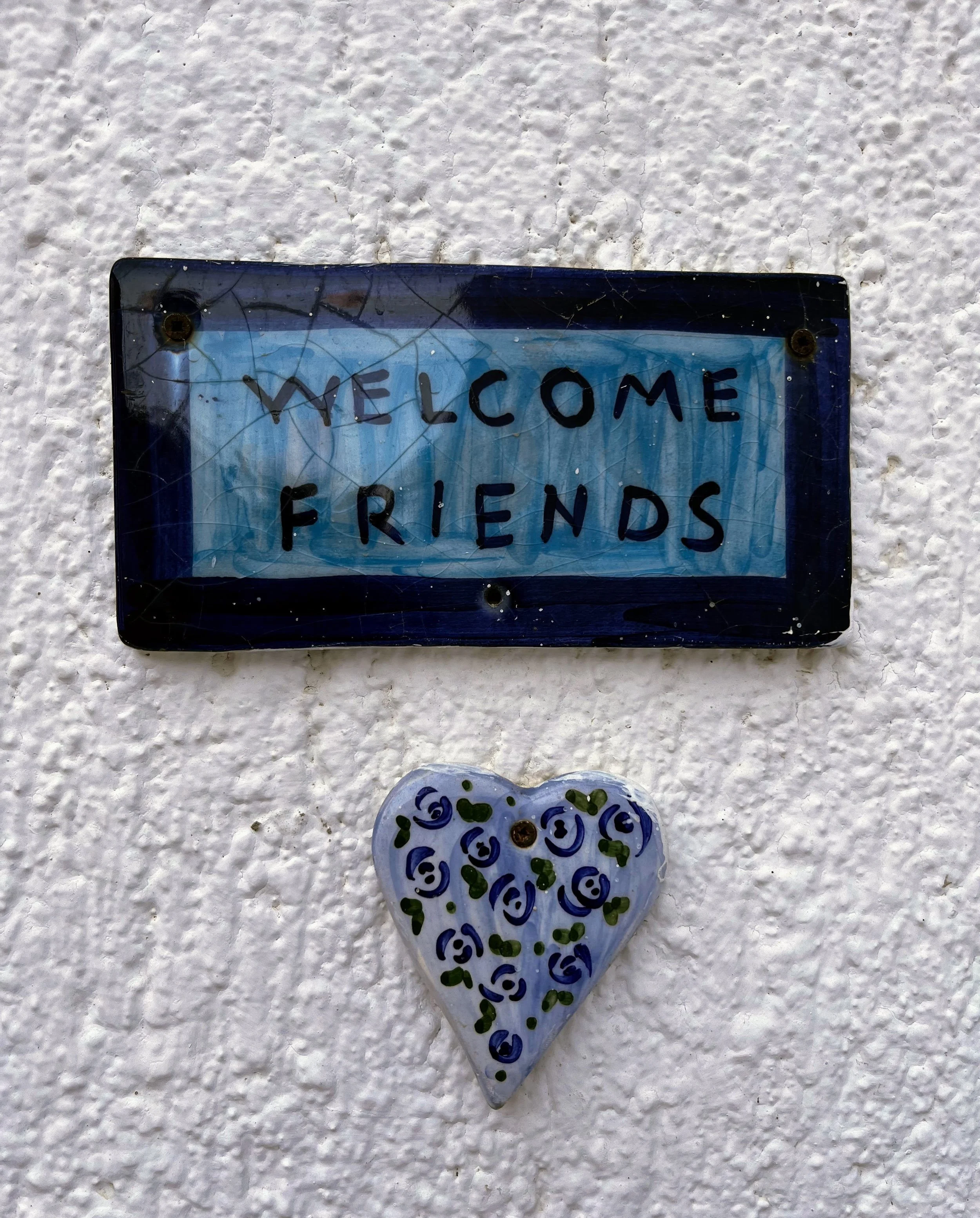A decorative sign on a white textured wall that reads 'Welcome Friends' and a small heart-shaped ornament with blue and green swirl patterns hanging below it.