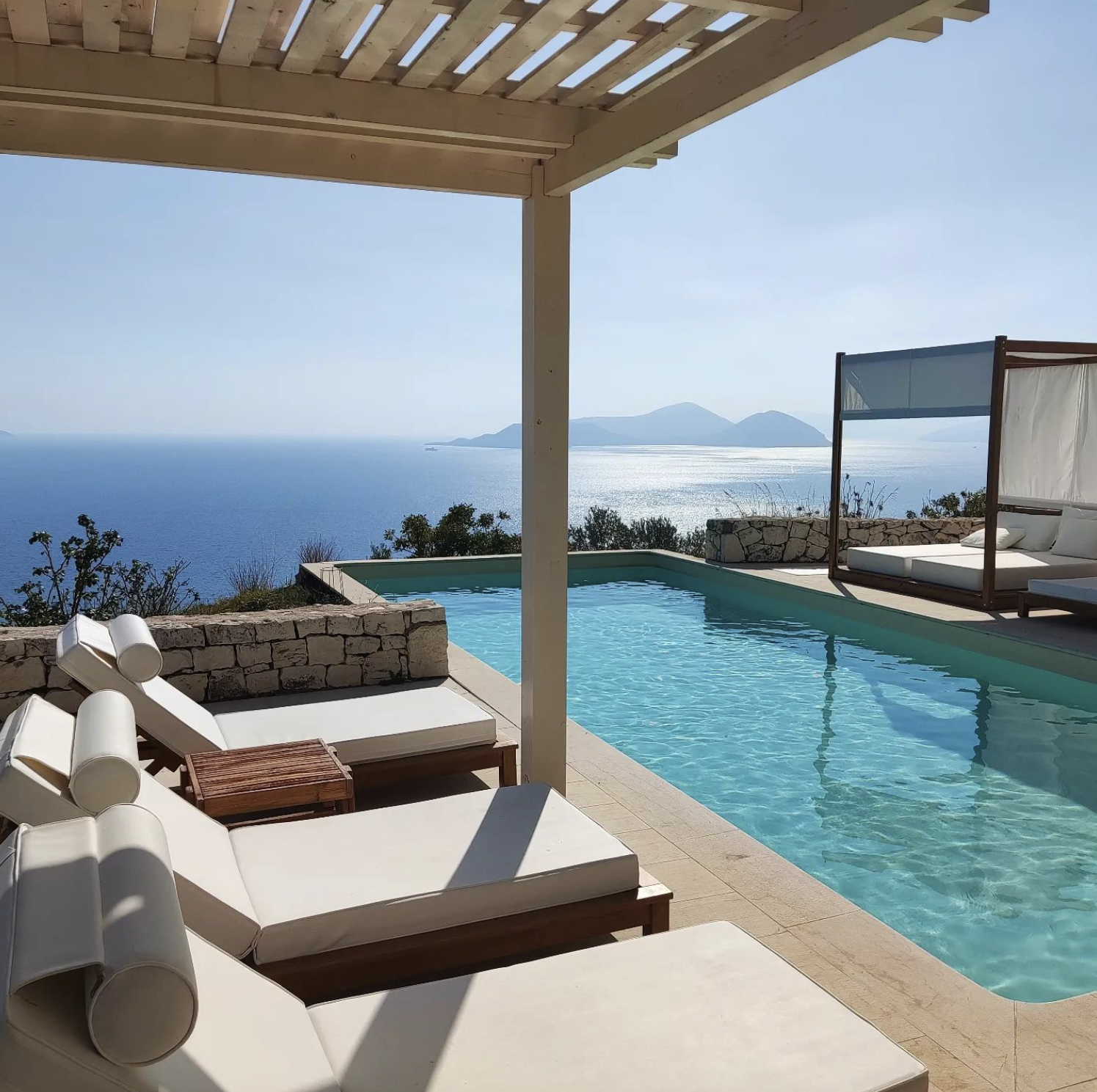 View of a luxurious infinity pool on a terrace overlooking the ocean with distant islands, cabanas, and white lounge chairs under a pergola.