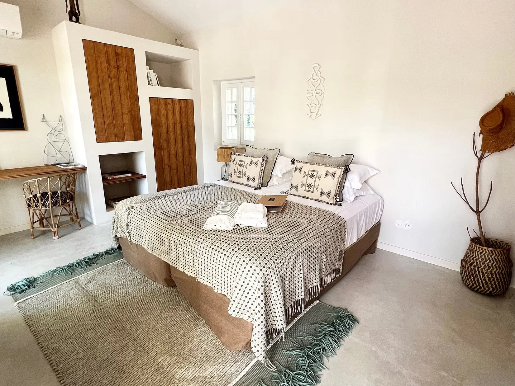 A bedroom with a bed featuring pillows and a beige and black checkered blanket. Next to the bed, a small nightstand with a lamp. To the right, a tall branch in a patterned basket. To the left, a corner with a wooden desk and chair, and a built-in white and brown cabinetry unit. A window lets in natural light. A beige rug covers part of the light-colored floor.